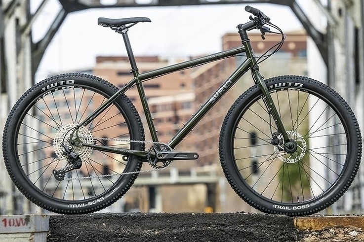 A glamour shot of the 2023 Surly Bridge Club. A touring/mountain/gravel bike. It’s a classic double-diamond shaped bike. The drive train is one cog on the front and 11 on the rear wheel. This one is a dark green colour with knobbly gravel tyres. 