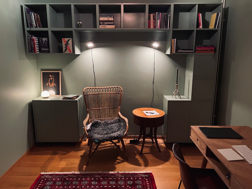 A cozy room with Xanadu green walls, one are mostly occupied with cube shaped cabinets in different sizes. Someone have started filling the shelves with books, but there are still a lot of ones. A large, comfortable-looking armchair invites you to relax and maybe read some.