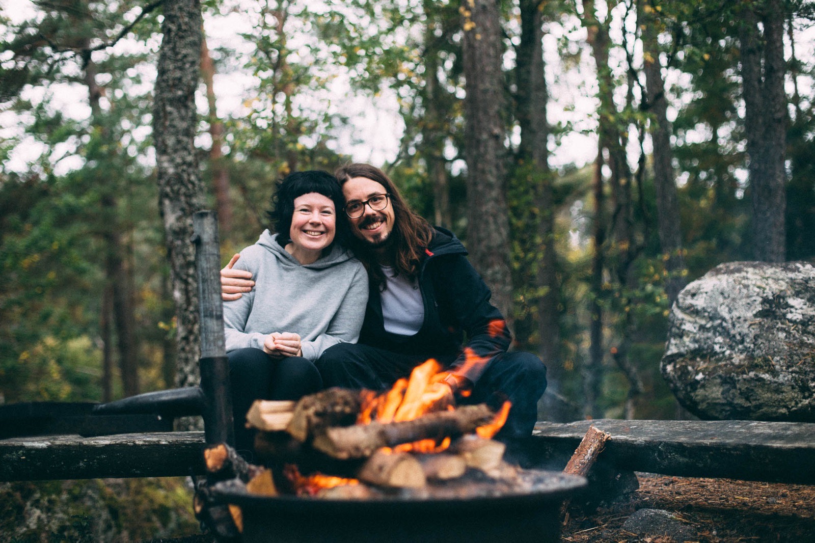 A smiling couple in a forest. Resting on a log close to a campfire.