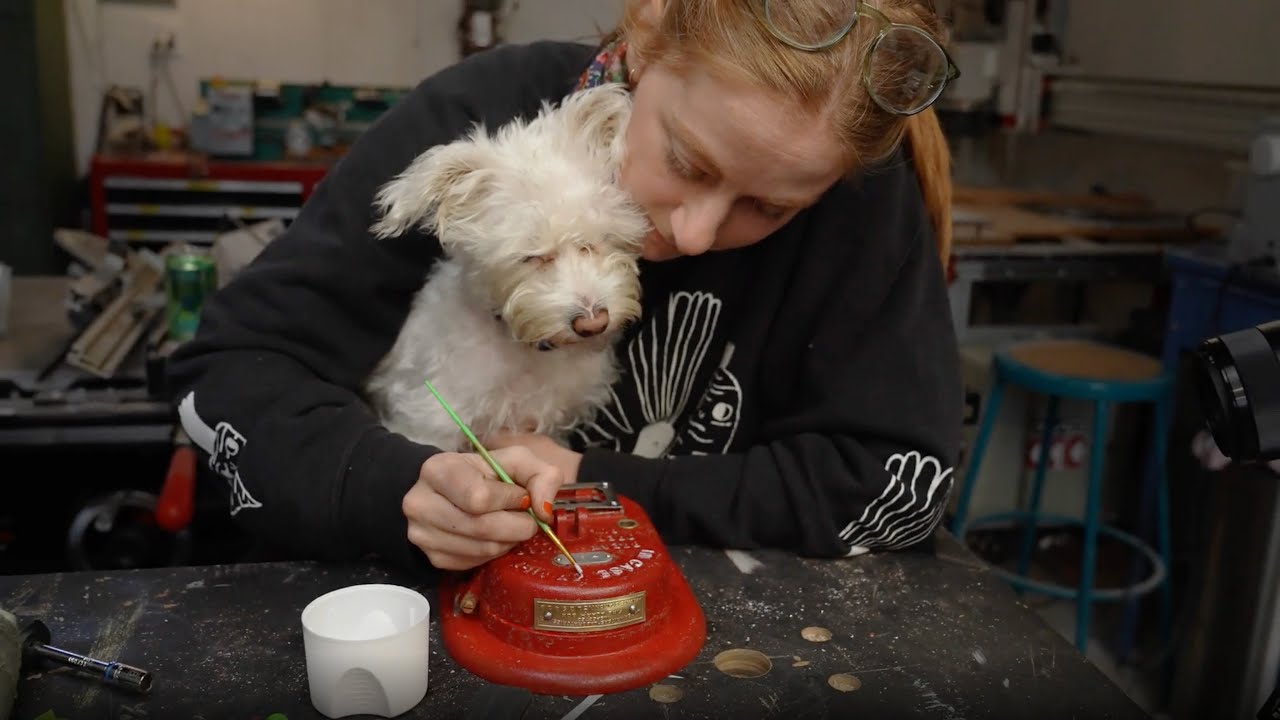 A woman sits at a desk, painting the letters of an old fire alarm. There’s a dog in her lap following the paintbrush’s motions.