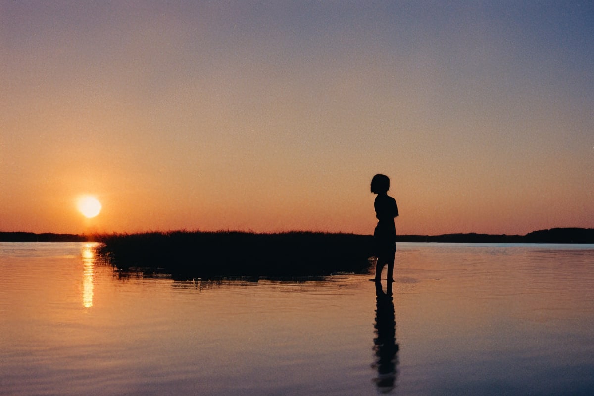 Silhouette of a woman immersed in calm, shallow waters, gazing at the fiery hues of the setting sun.