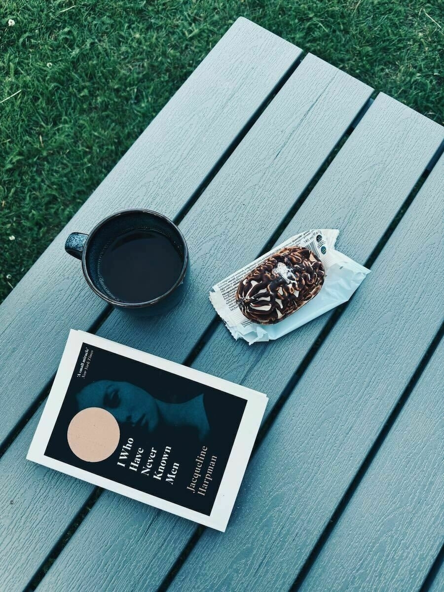 Overhead view of a book, a coffee mug, and an ice cream on a wooden table, with grass visible in the background. The book cover of I Who Have Never Known Men by Jacqueline Harpman features a silhouette of a woman with an orange, circular shape partially covering her face.