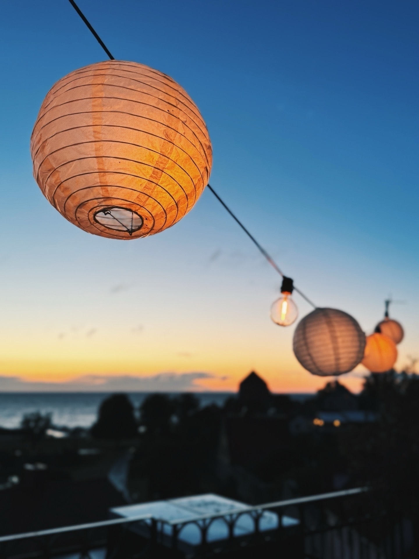 A close-up of lit paper lanterns hanging on a string, set against a twilight sky with a silhouetted landscape and the Baltic Sea in the distance.