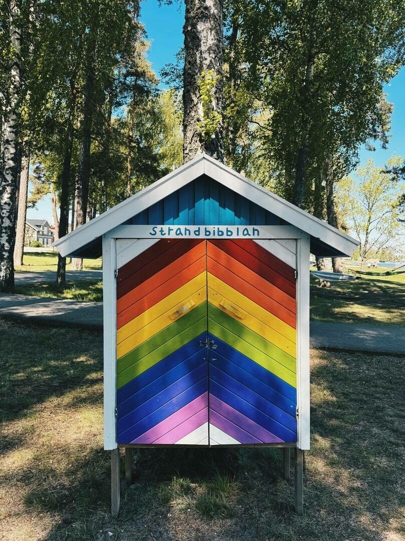 A colorful wooden shed painted with rainbow stripes between two birch trees. The text Strandbibblan (the Beach Library) is written across the top.