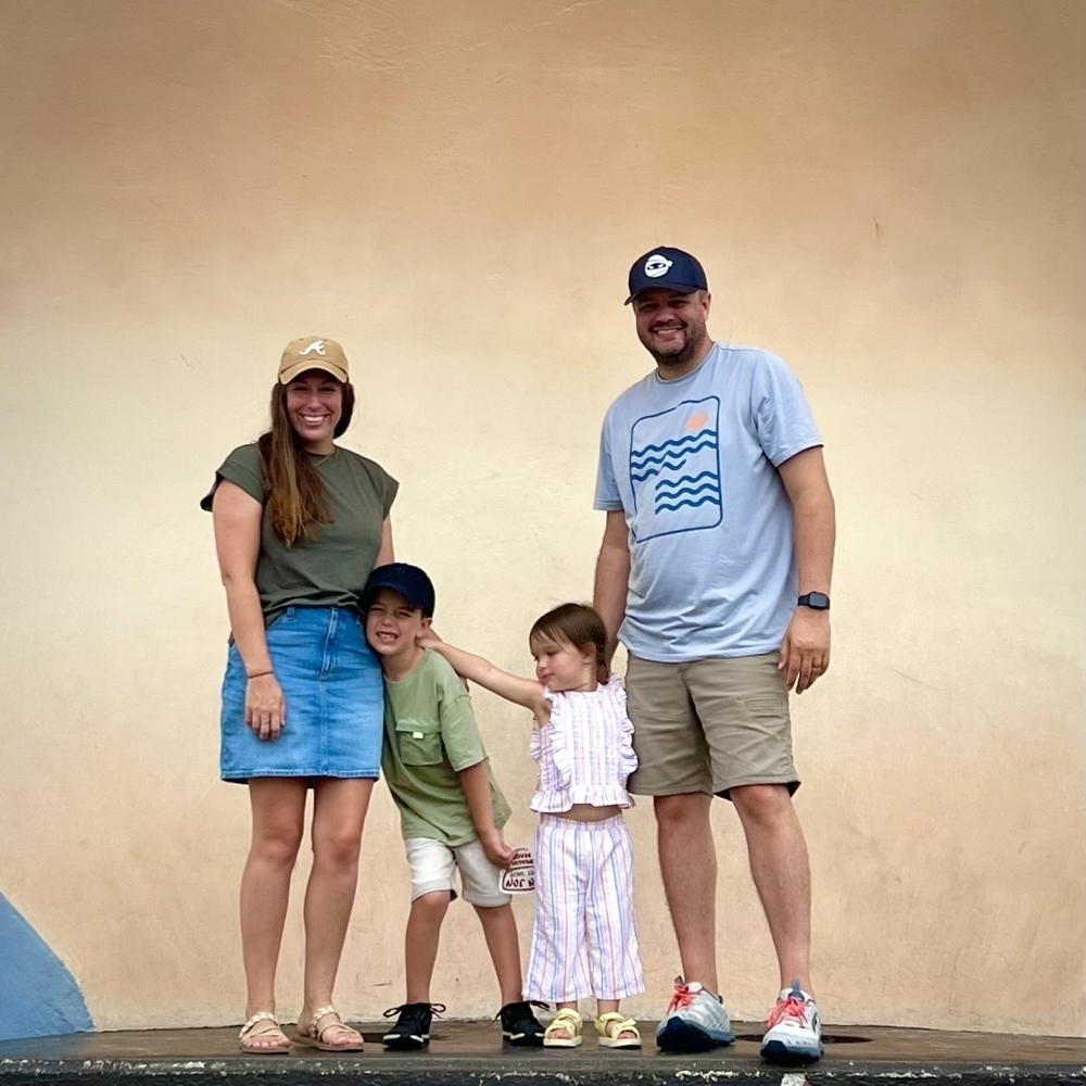 A family of four stands smiling against a plain backdrop, with the children playfully posing between the parents.