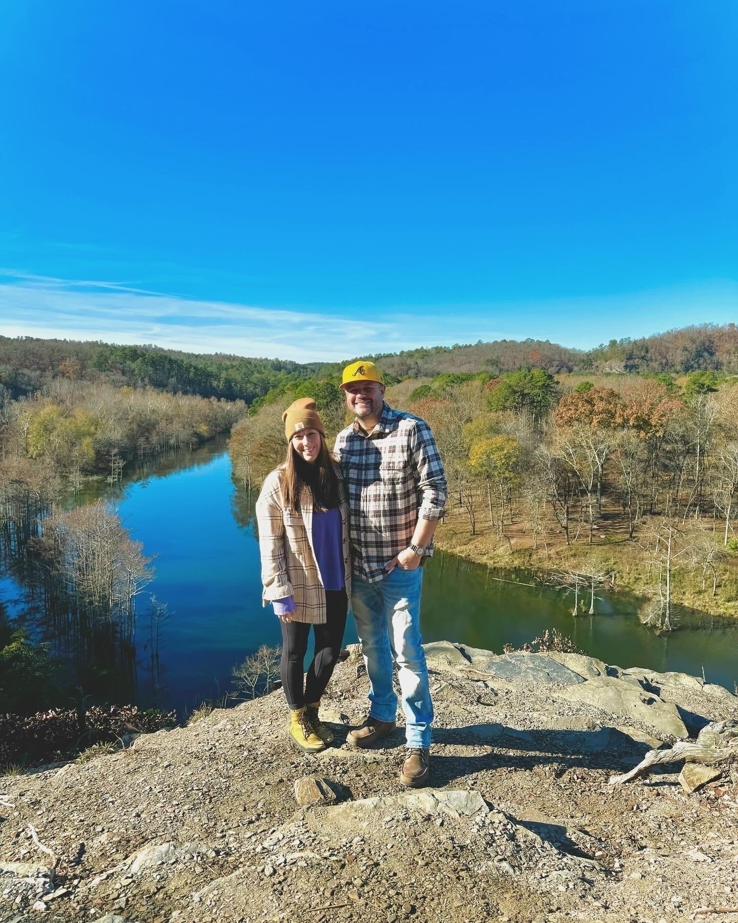 A man and woman in casual outdoor attire are standing on a rocky ledge overlooking a scenic landscape with a river and forested area.