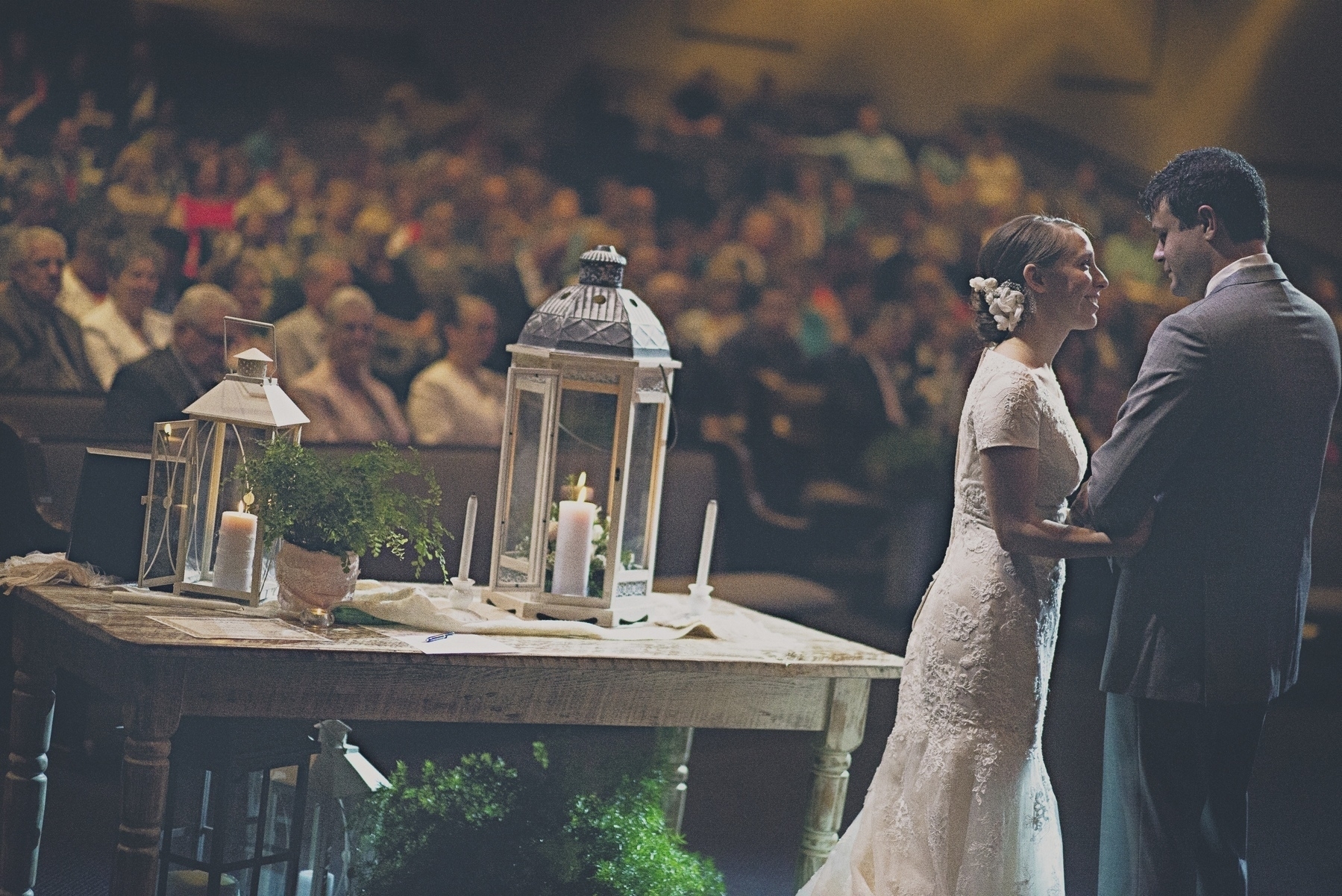 A couple is standing in front of a beautifully decorated table with lanterns and candles, holding hands and gazing at each other, while an audience watches.