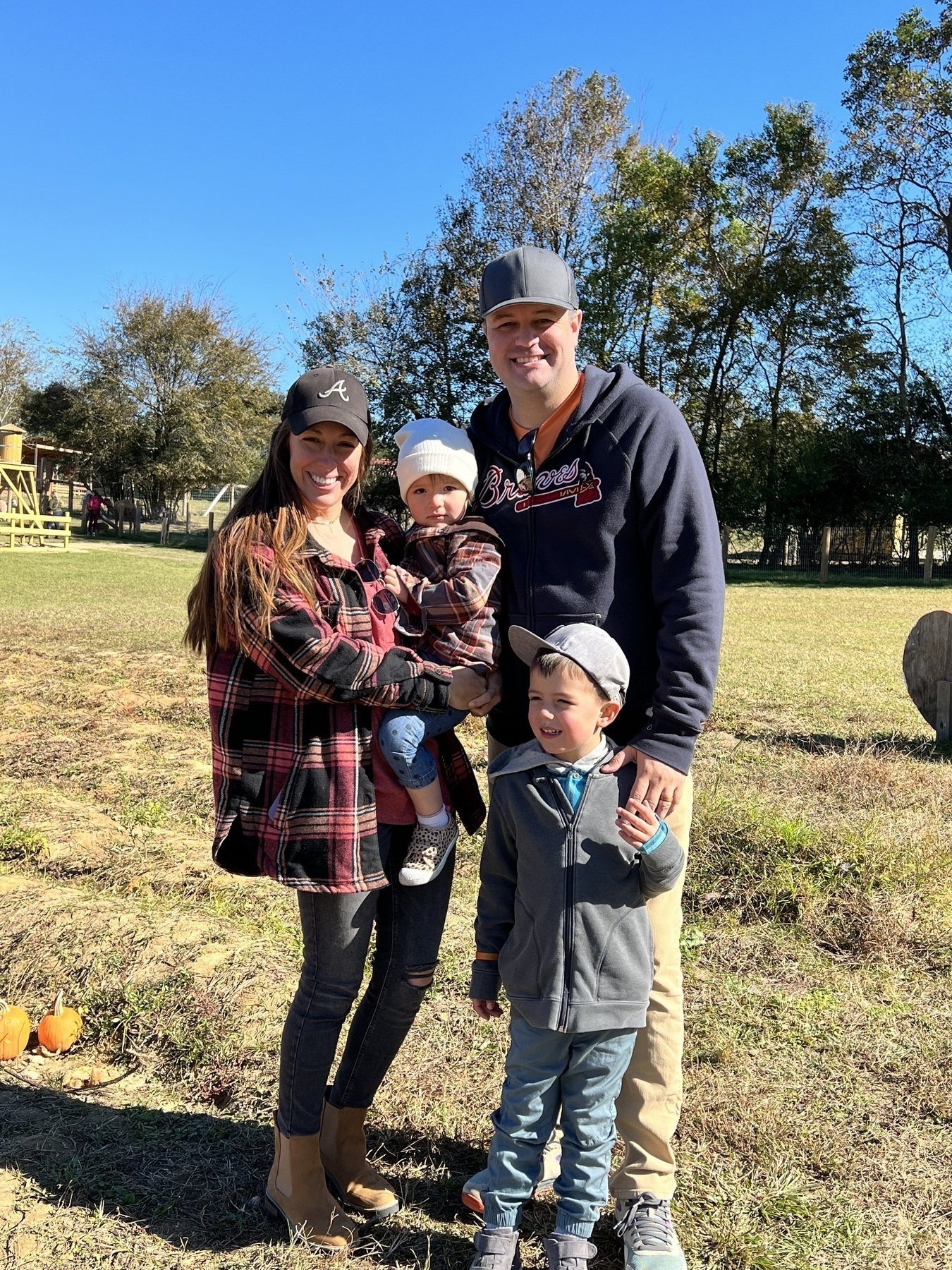 A family of four is smiling outdoors in a grassy area with trees and a clear blue sky in the background.