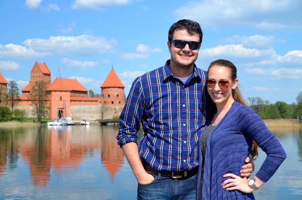 A smiling couple wearing sunglasses stands in front of a scenic view featuring an old brick castle by a body of water under a clear blue sky.