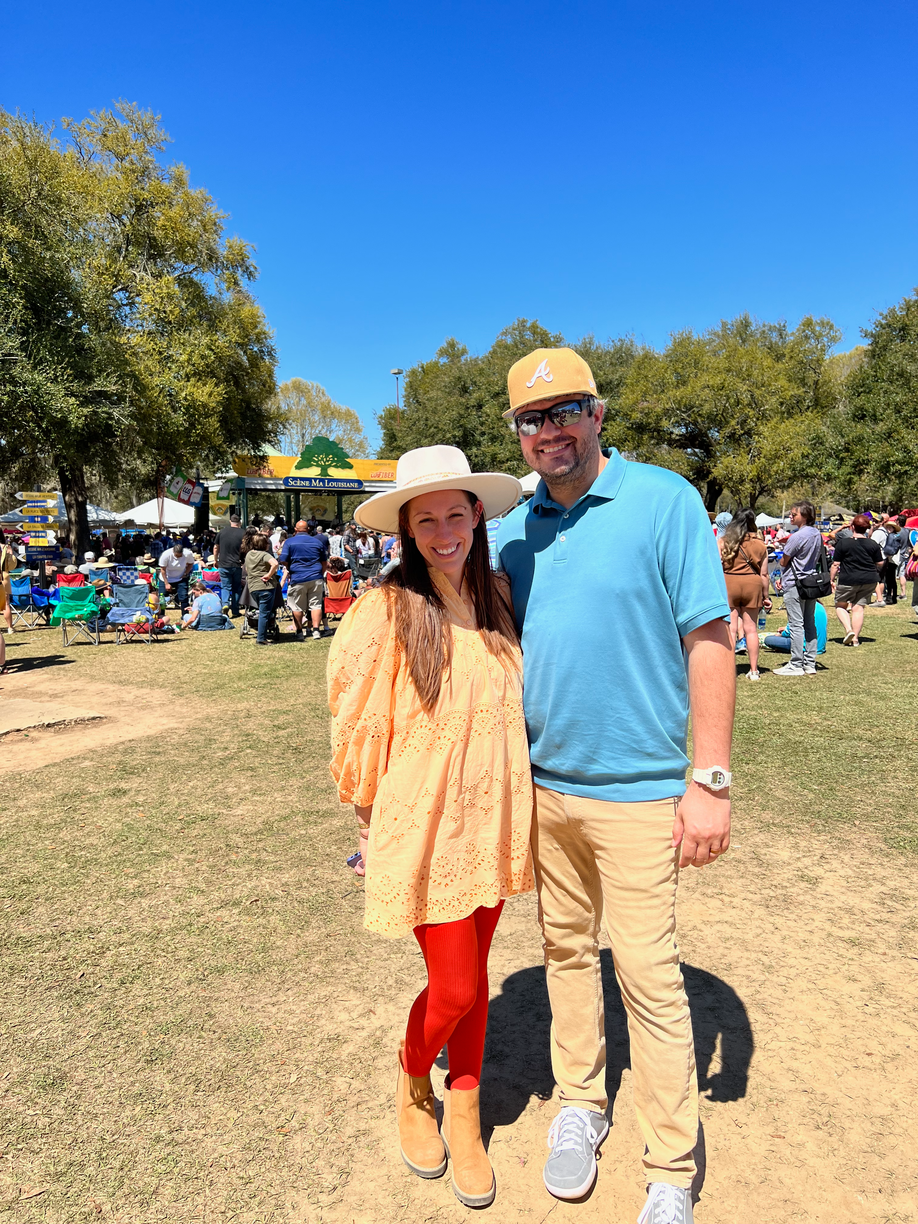 A smiling couple poses outdoors at a sunny event, surrounded by trees and people.