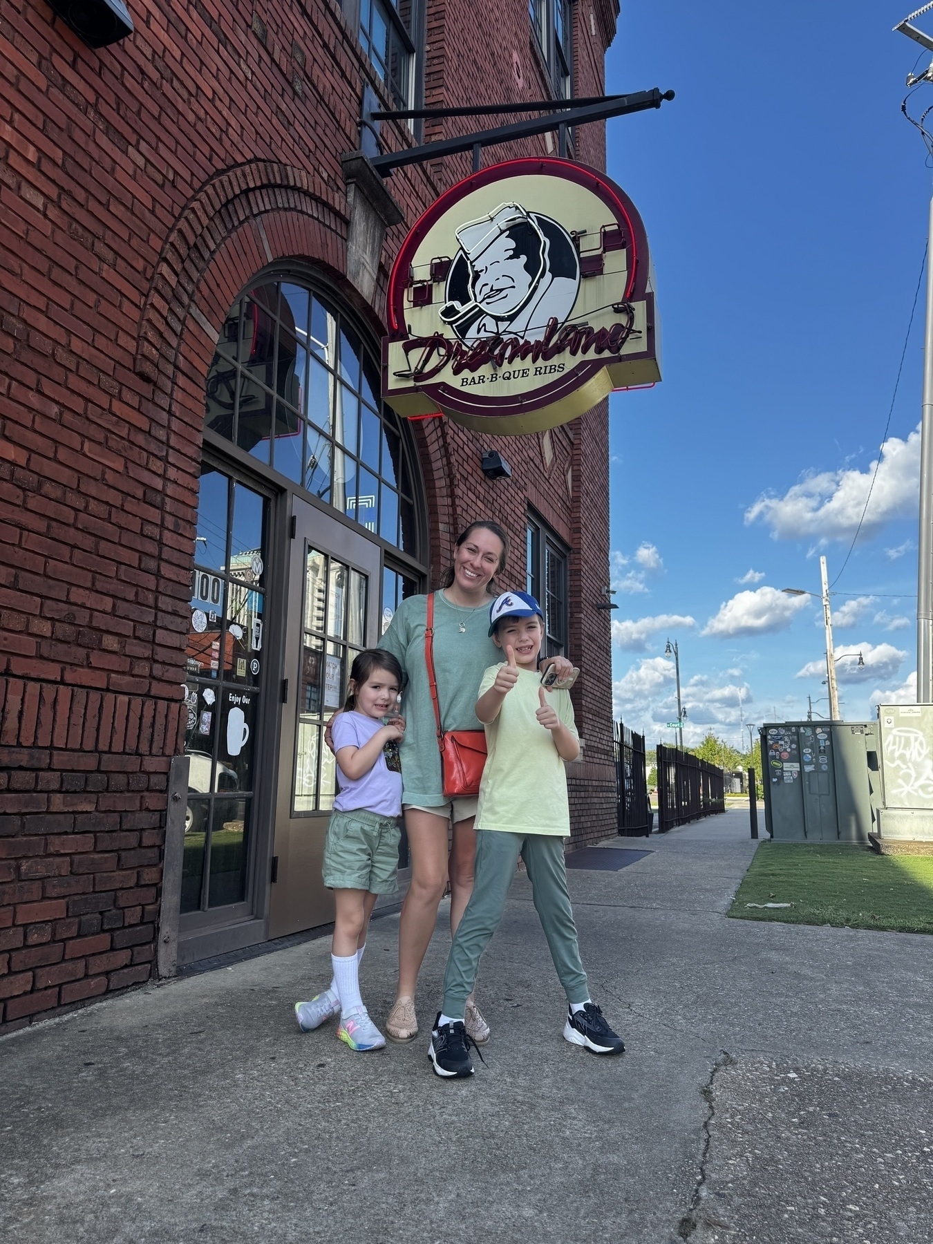 A woman and two children are standing in front of a brick building with a Dreamland BBQ sign above them.