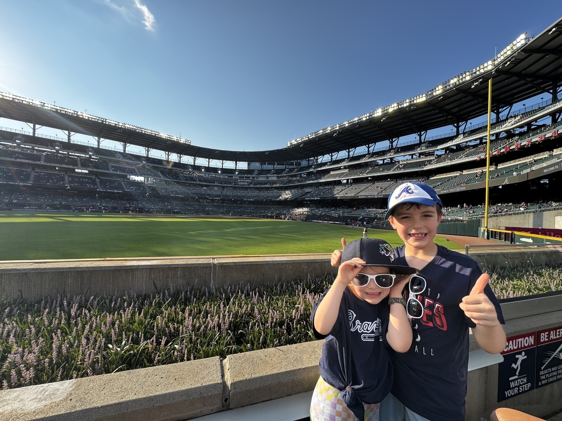 Two children, both wearing baseball caps and t-shirts, stand smiling and giving thumbs up near the stands of a baseball stadium.