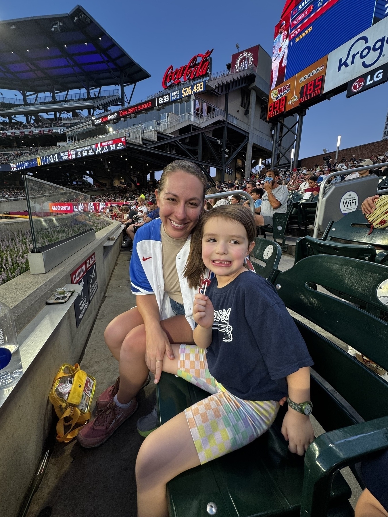 A woman and a young girl are sitting in a stadium, both smiling at the camera with a crowd of spectators and various advertisements visible in the background.