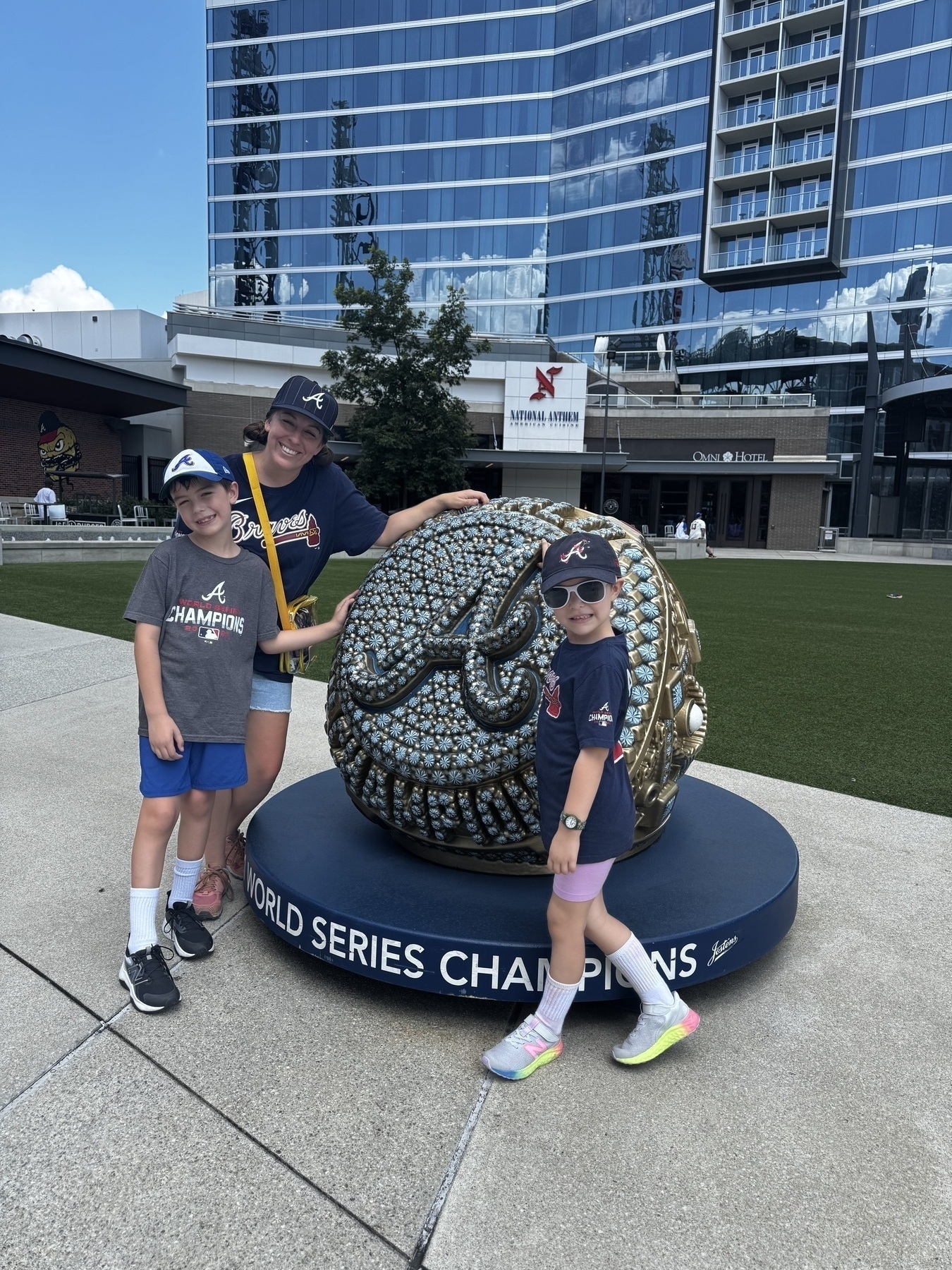 Three people wearing Braves gear stand in front of a large World Series Champions baseball sculpture near a modern building.