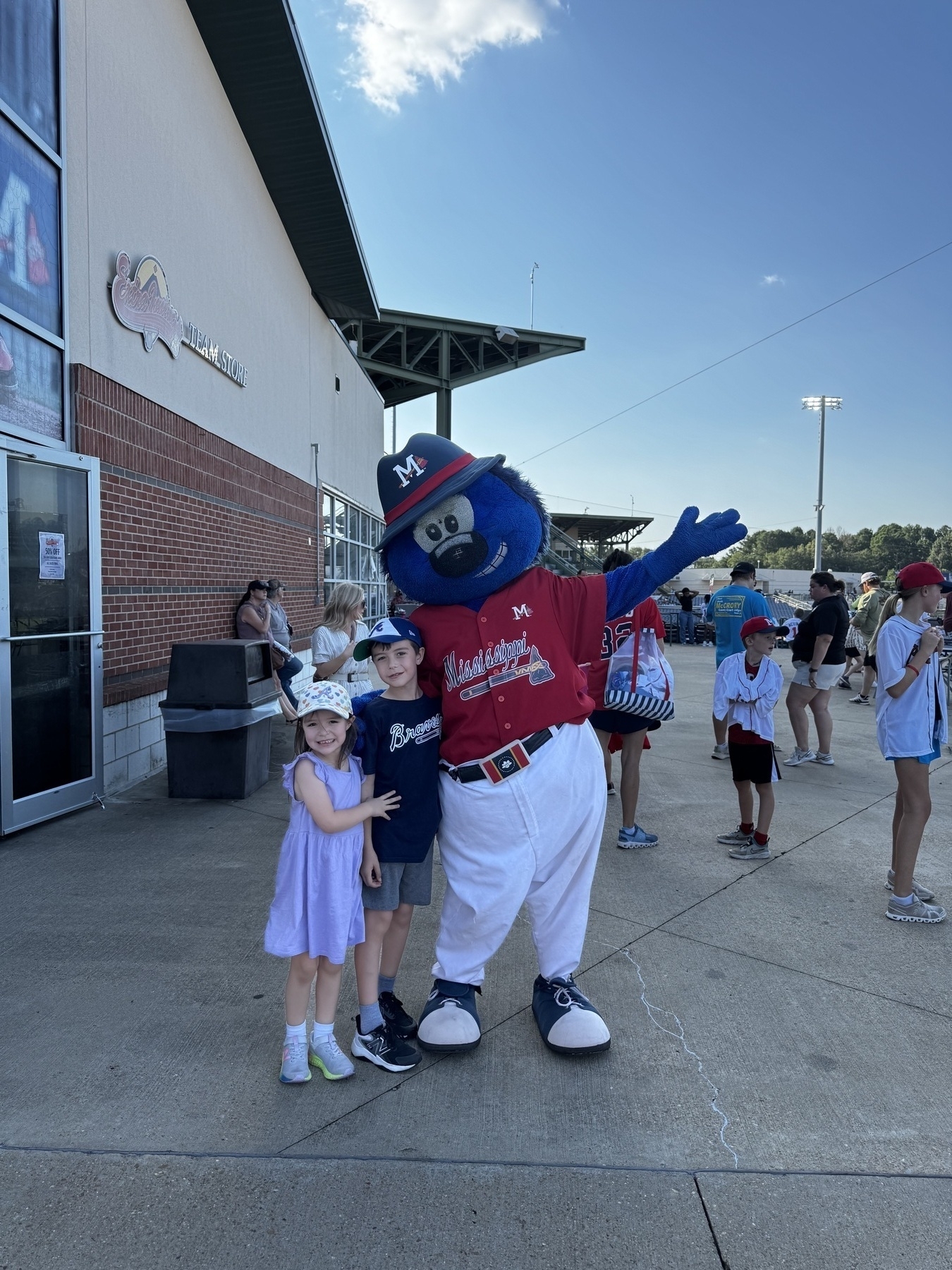 A blue mascot in a baseball uniform poses with two children in front of a stadium building.