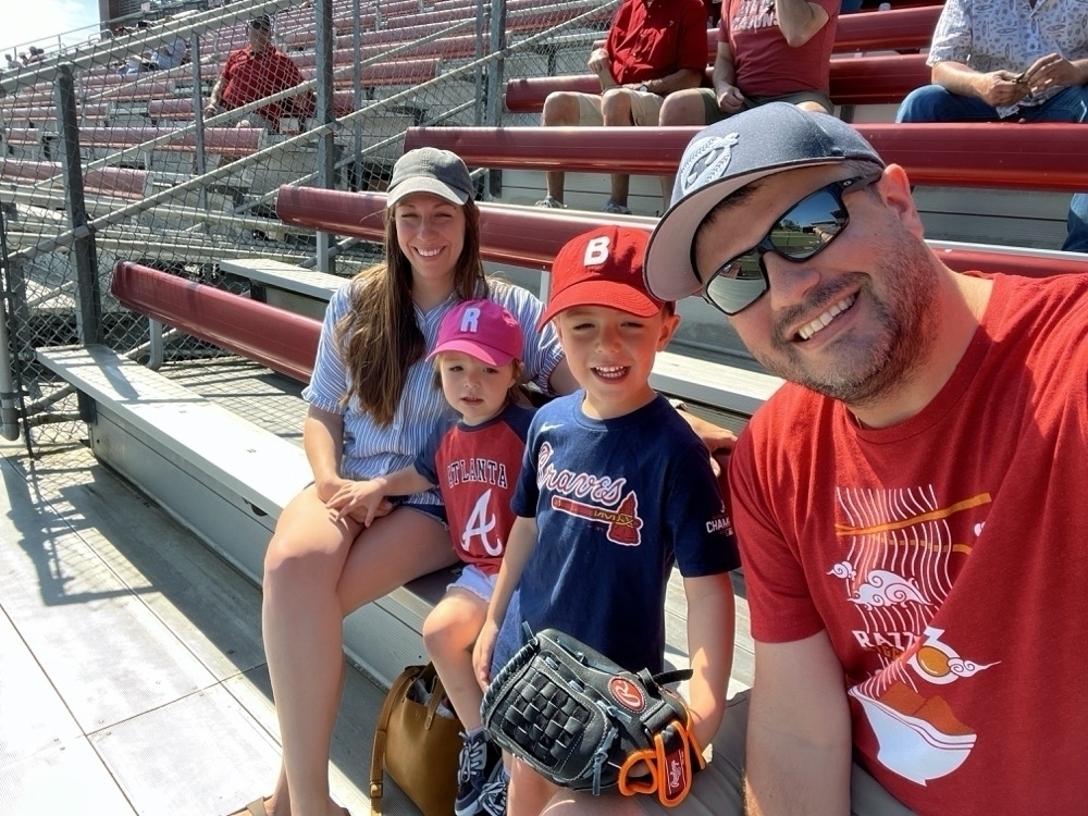 A group of four—two adults and two children—are sitting on bleachers, smiling for a selfie with a baseball field in the background, and they appear to be enjoying a sunny day at a baseball game.