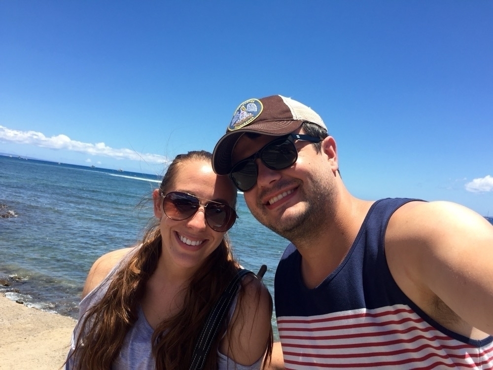 A smiling man and woman are taking a selfie on a sunny day with a clear blue sky and the ocean in the background.