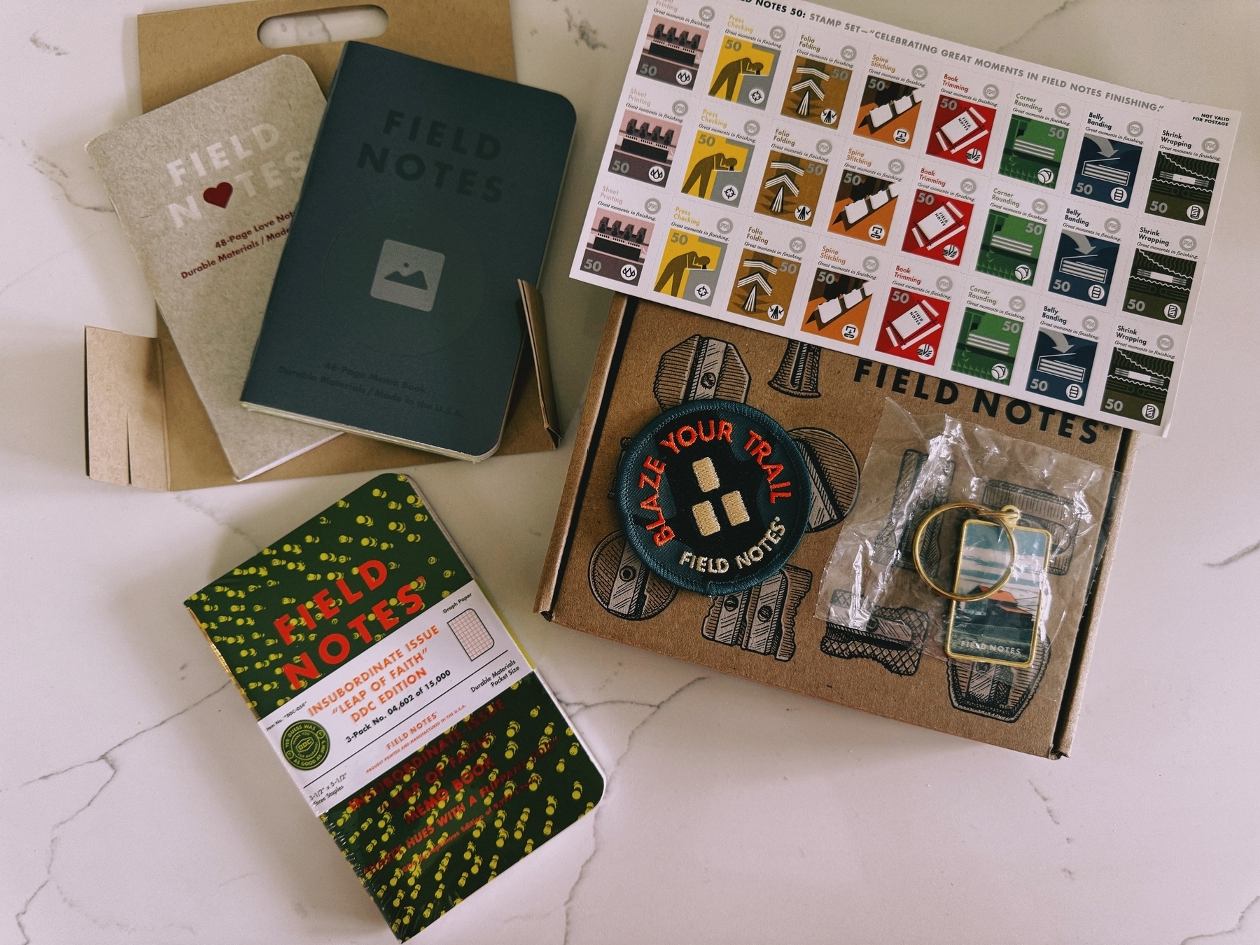 A collection of Field Notes notebooks, a chart with colored tabs, a "Blaze Your Trail" patch, and a keychain are arranged on a marble surface.