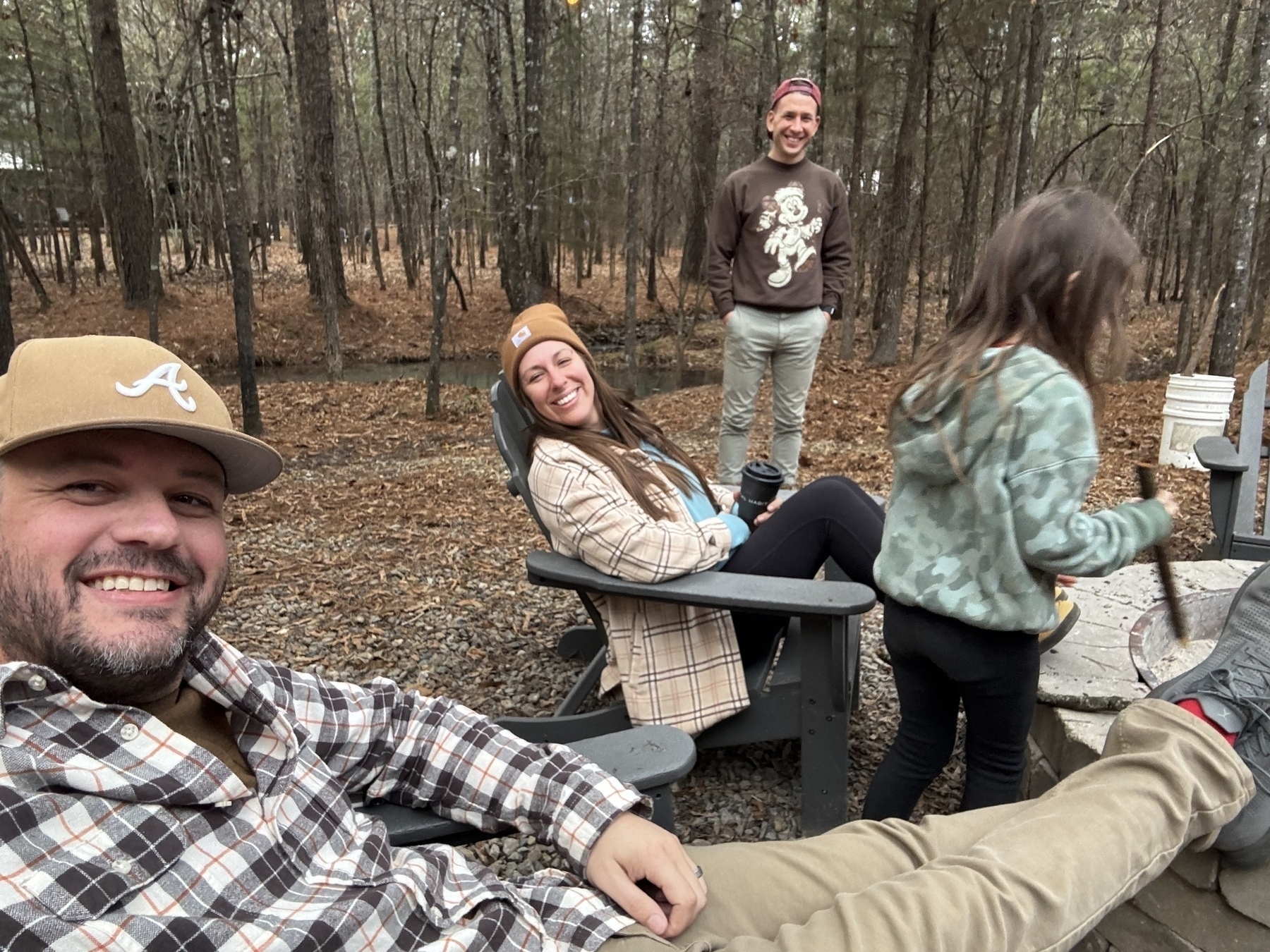 Four people are relaxing outdoors in a wooded area, wearing casual attire and enjoying each other's company.