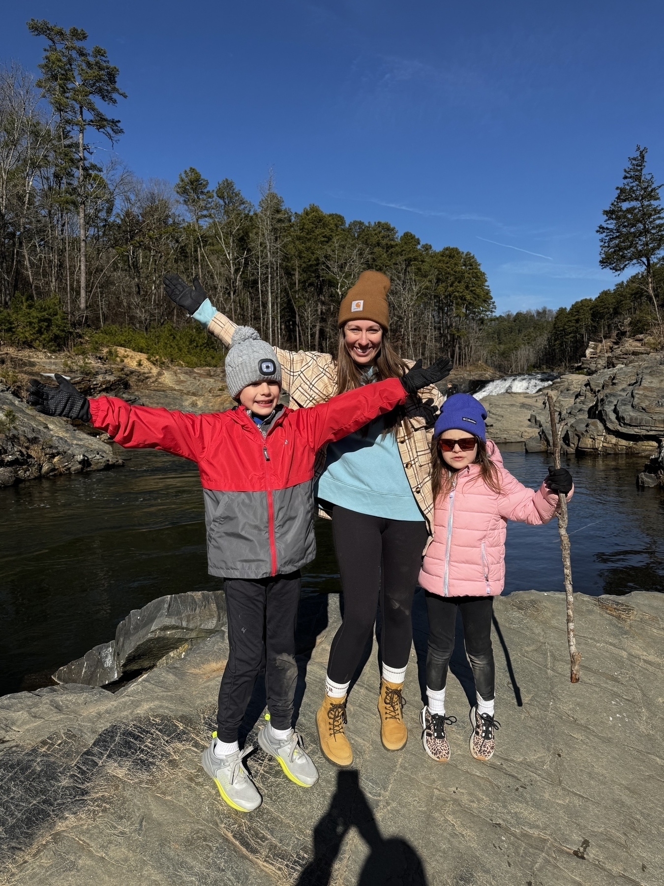 A woman and two children, all wearing winter clothing, pose cheerfully on a rocky area near a river.