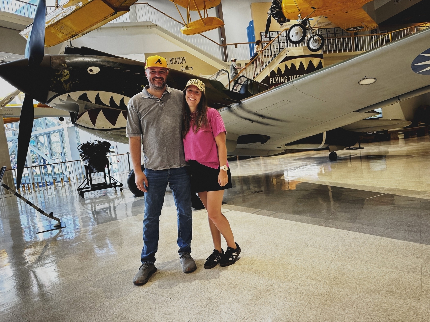 A man and a woman wearing yellow caps are posing in front of a vintage aircraft at a museum.