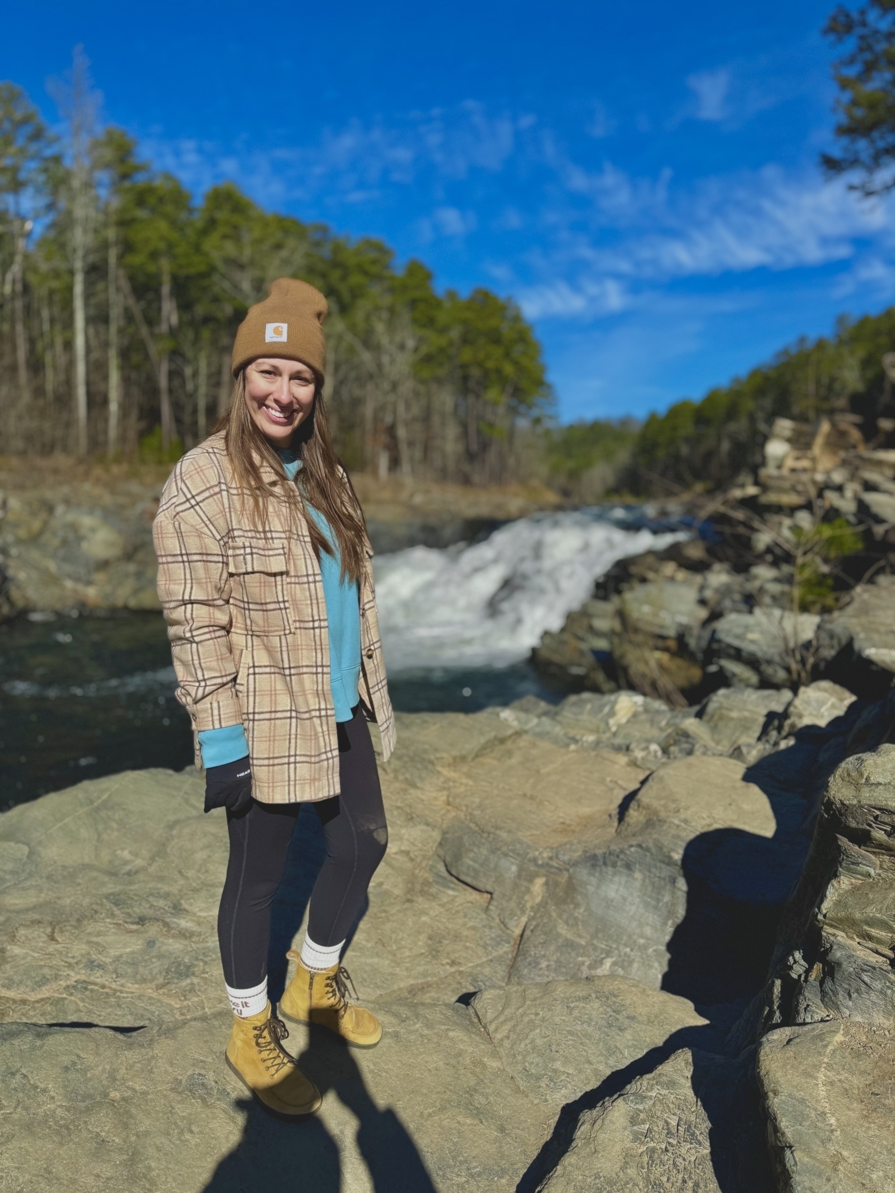 A person wearing a plaid jacket and a beanie stands on rocks near a river with a waterfall in a wooded area.