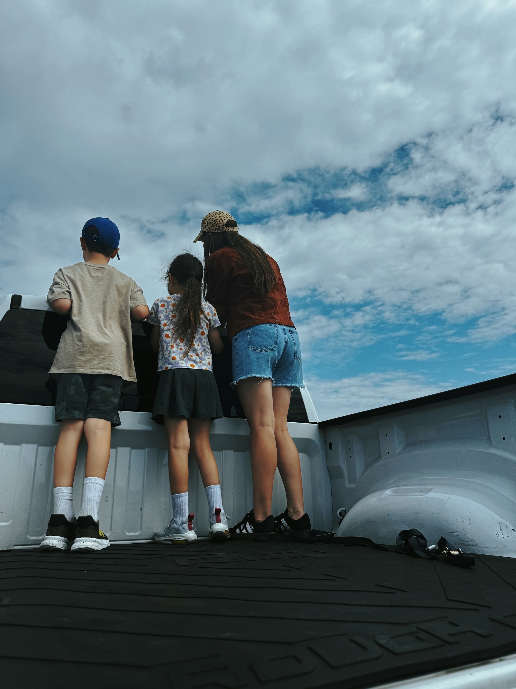Three people stand in the back of a truck looking at planes under a partly cloudy sky.