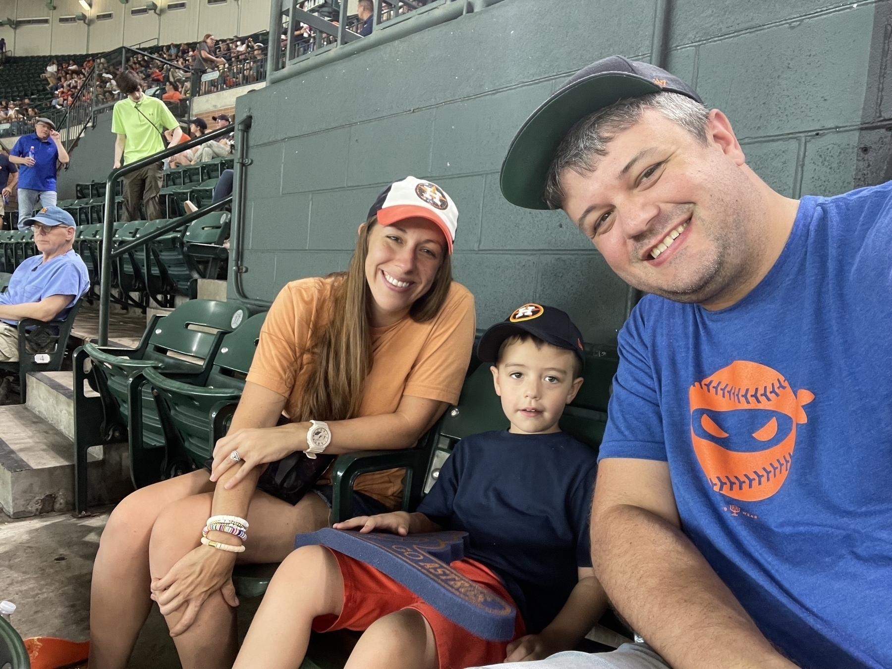 A smiling family of three is sitting in stadium seats, wearing casual clothes and caps.