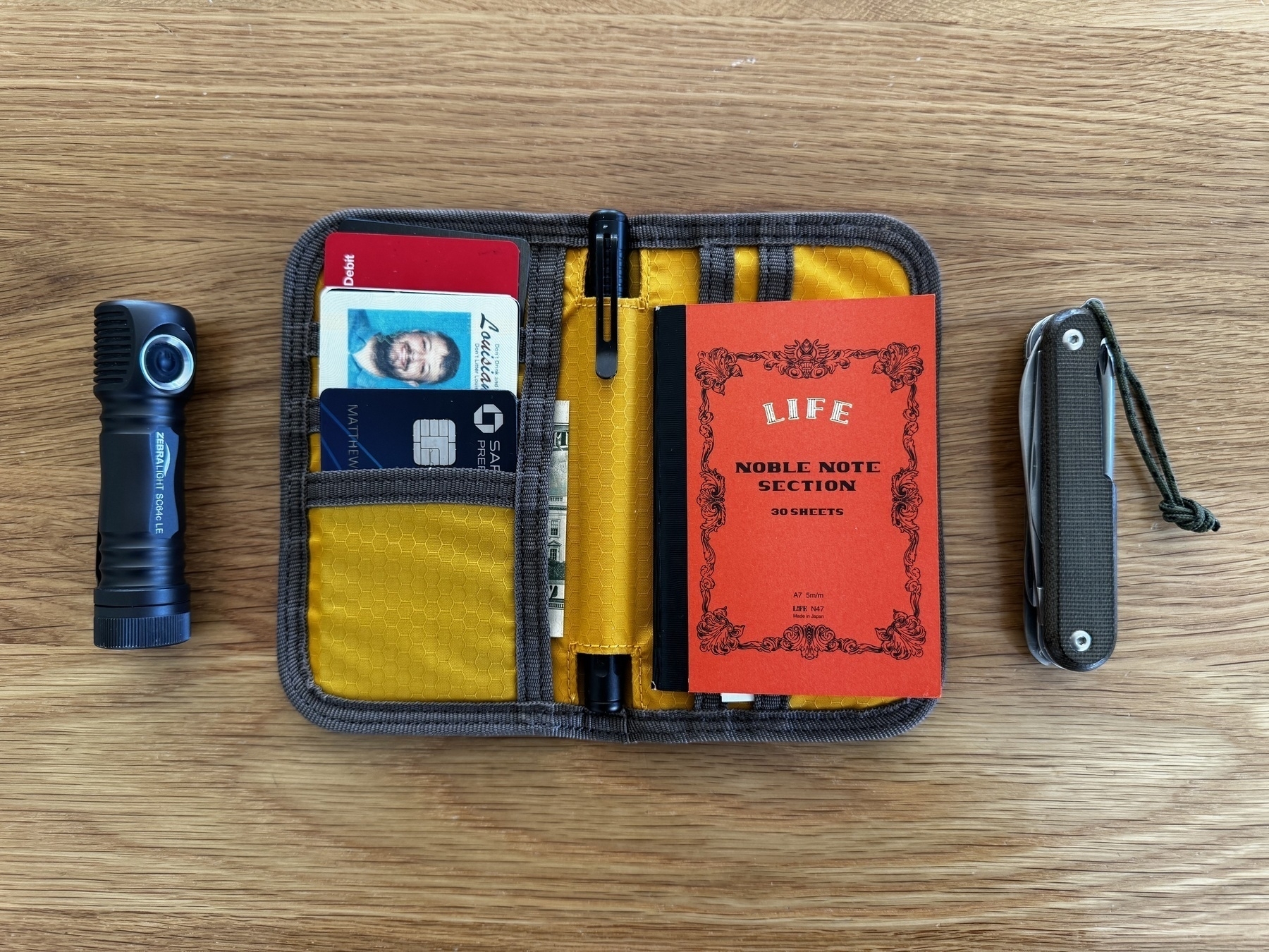 A neatly arranged pocket organizer containing cards, a pen, and a red notebook is flanked by a flashlight on the left and a multitool on the right.