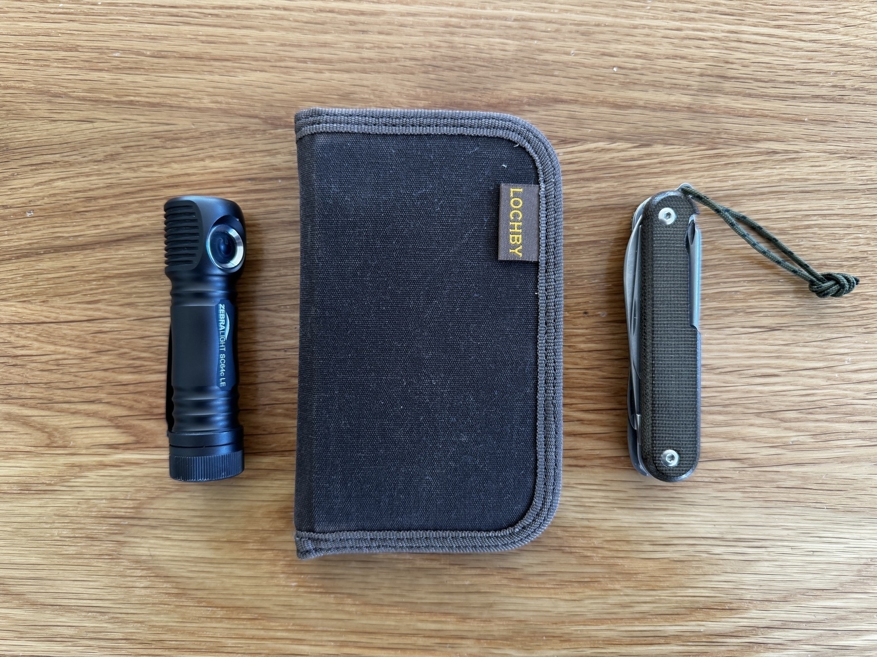 A small flashlight, a Lochby Sidekick, and a folding knife with a lanyard are laid out on a wooden surface.