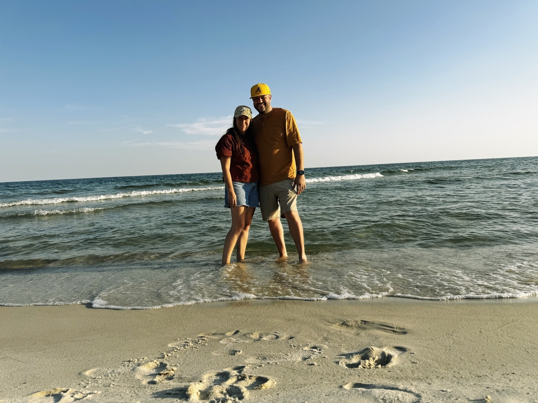 Auto-generated description: A couple stands on the beach with their feet in the surf, enjoying a sunny day.
