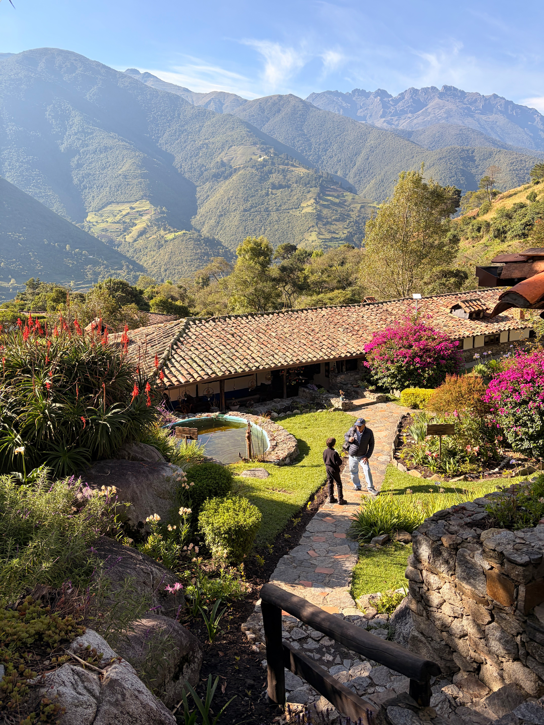 A serene mountainous landscape features a rustic stone path leading to a cottage surrounded by lush greenery and vibrant flowers.