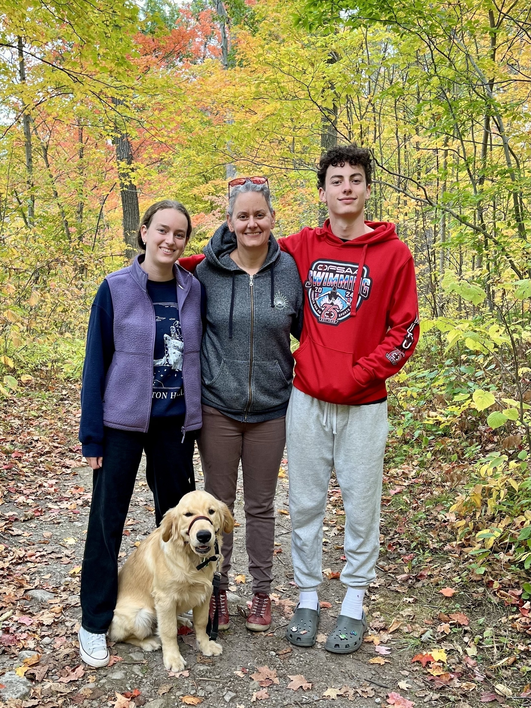 A group of three people and a dog are standing on a forest path surrounded by colorful autumn foliage.