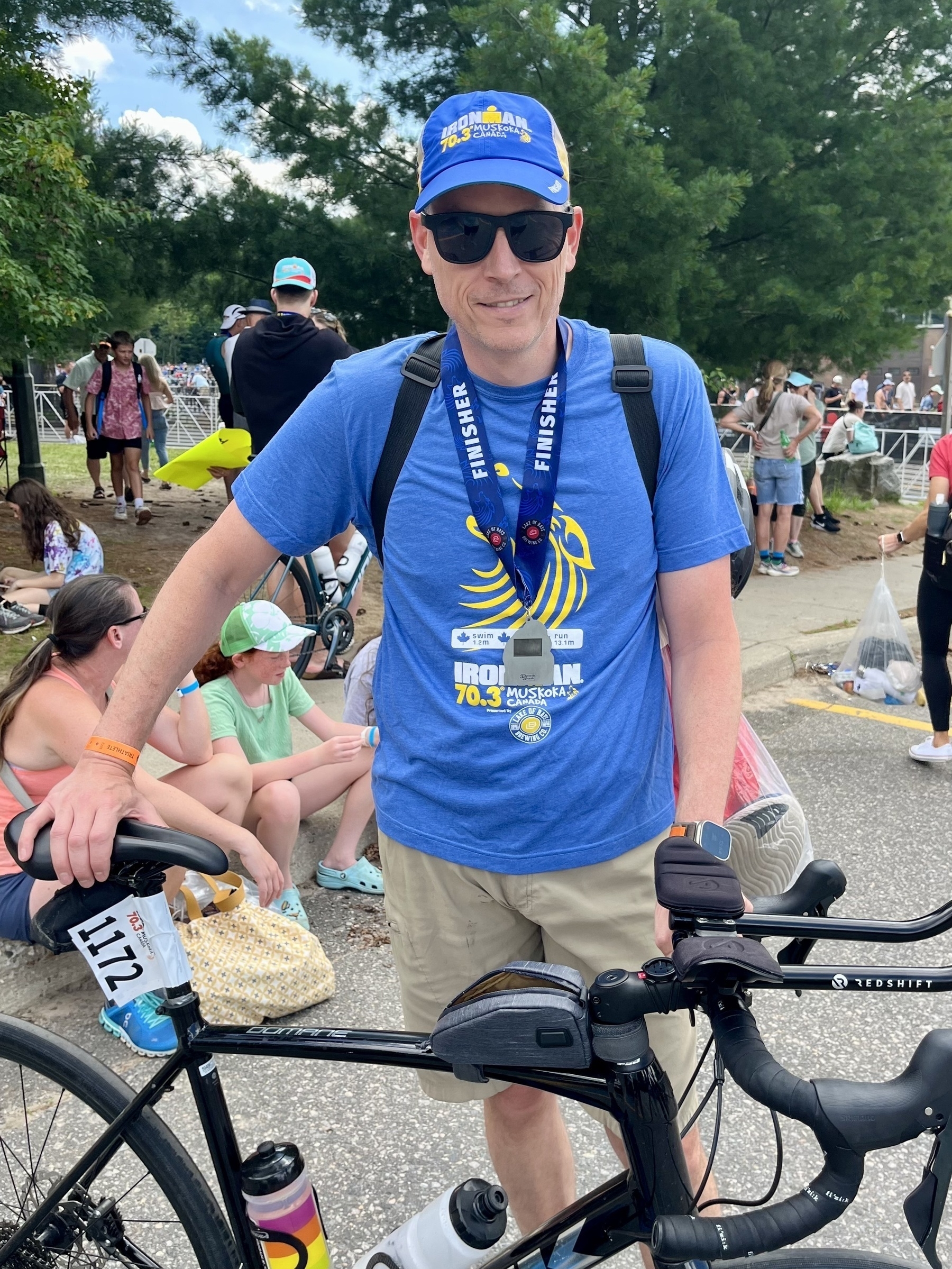 Auto-generated description: A man wearing blue finisher merchandise poses with his bicycle after completing an Ironman 70.3 triathlon event, with spectators in the background.