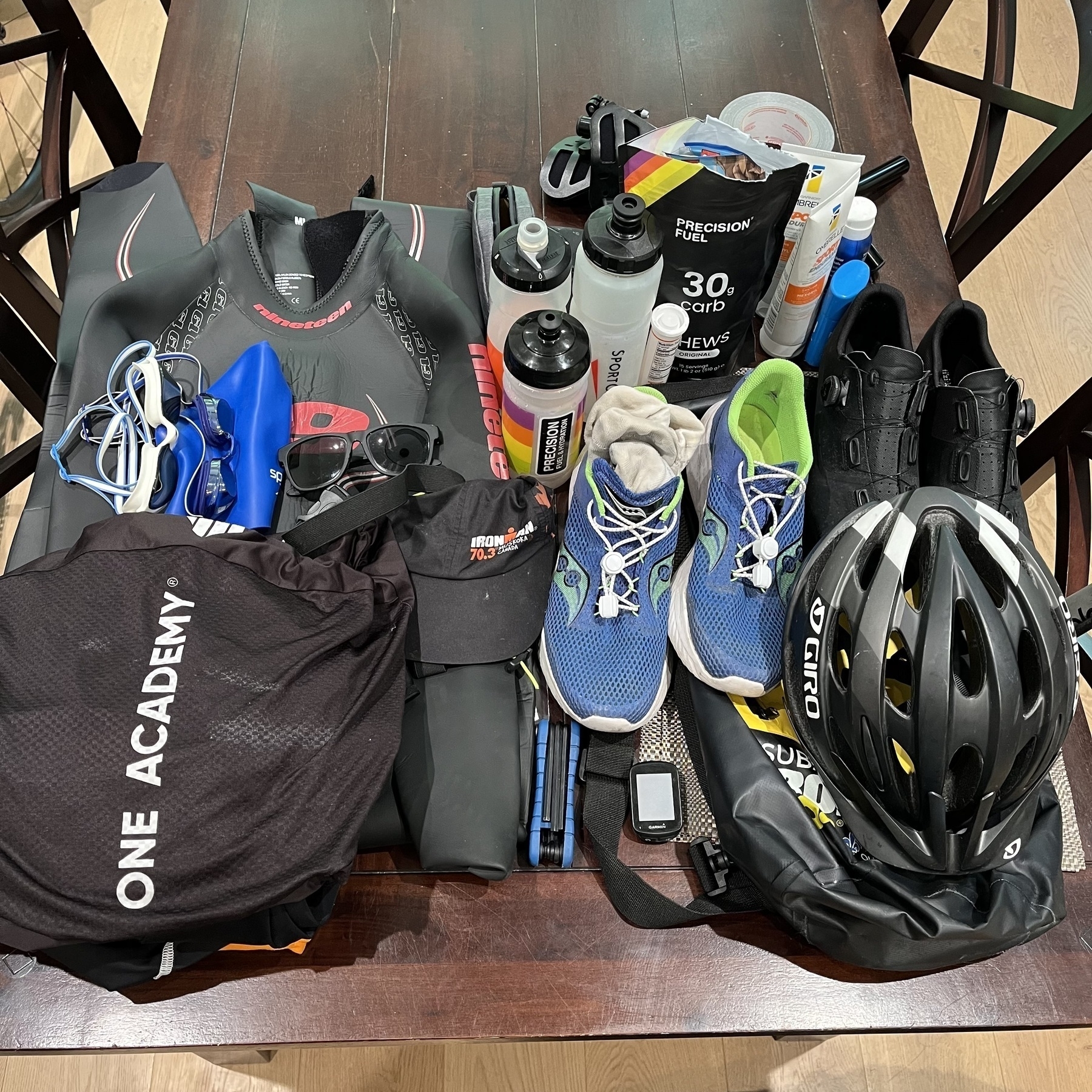Auto-generated description: A collection of triathlon gear arranged on a table, including a wetsuit, running shoes, helmet, water bottles, nutrition packs, a towel, and cycling shoes.