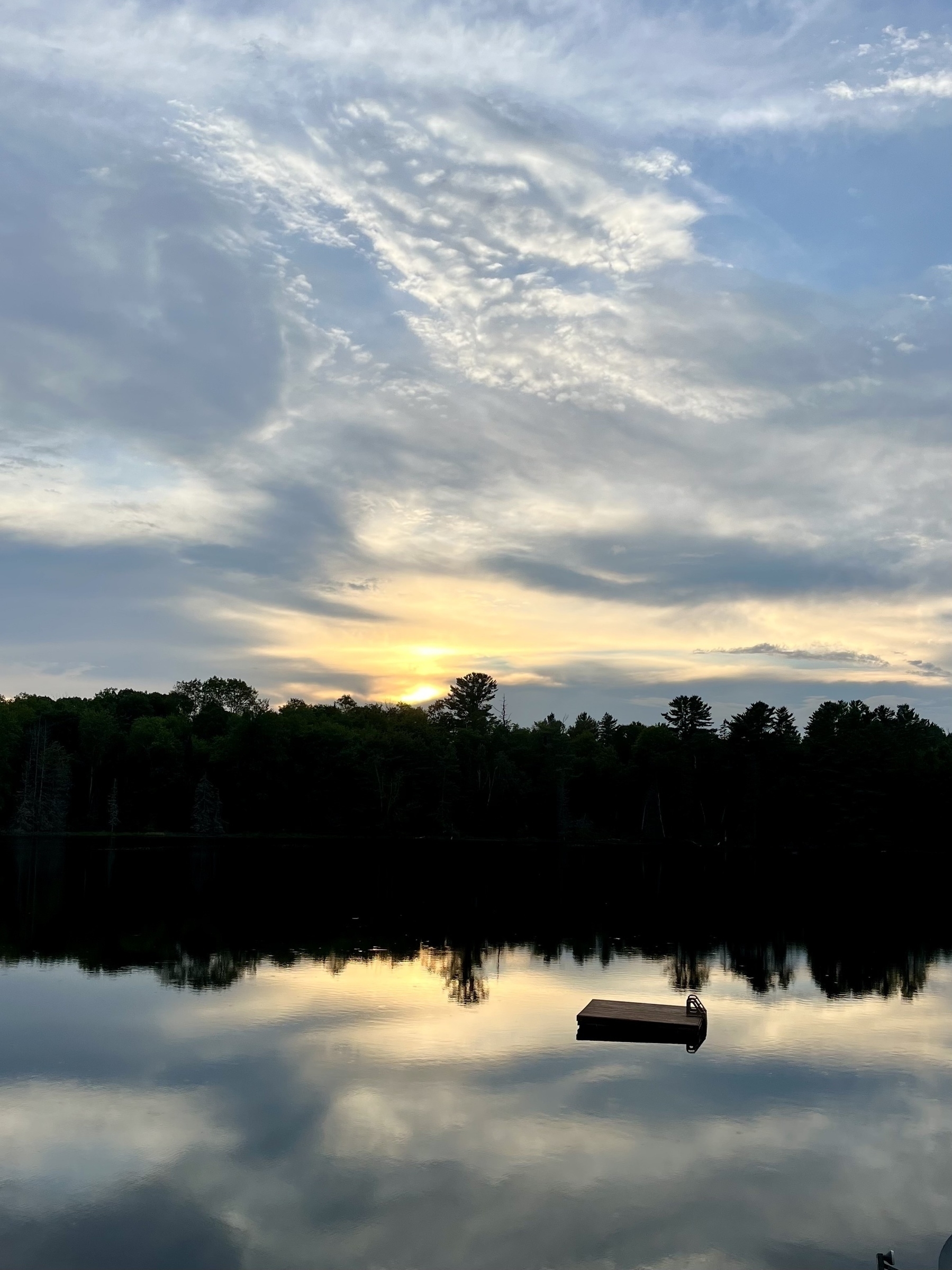 A tranquil lake reflects the colorful sunset sky and surrounding trees, with a small dock floating in the water.