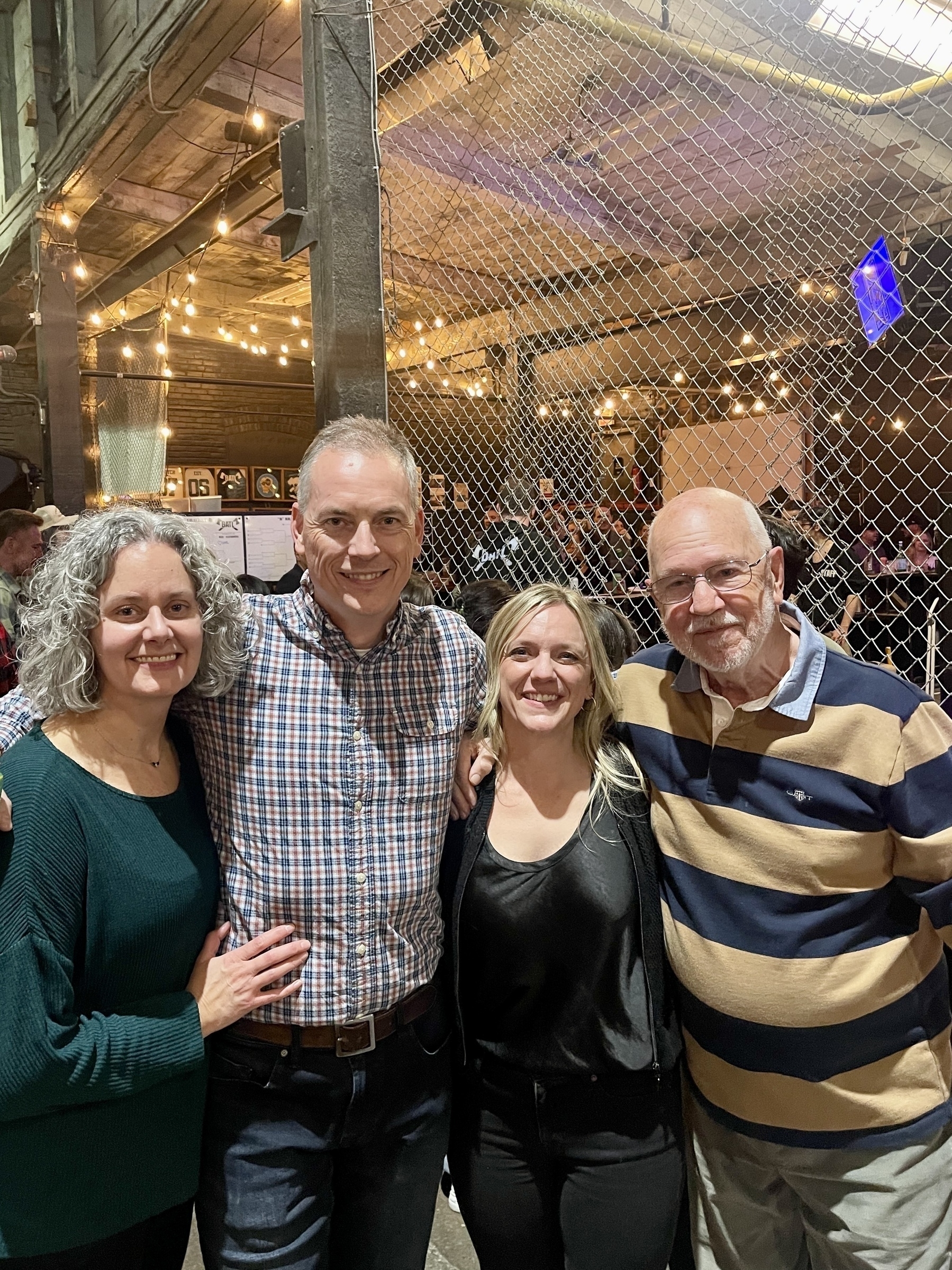 Four people are smiling and posing together indoors with string lights and a netted background.