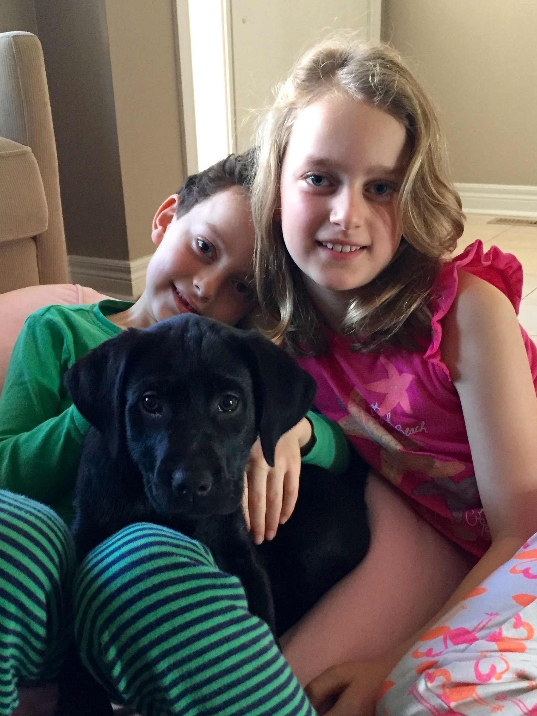 Two children are sitting together, smiling, and holding a black puppy.