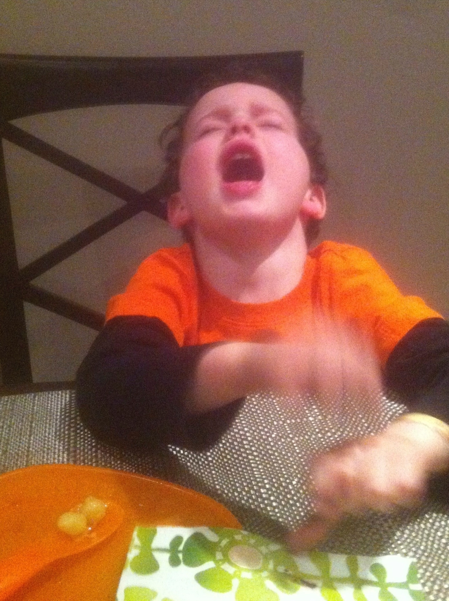A child is crying at a dining table with a blurred motion of hands, near an orange plate with food remnants.