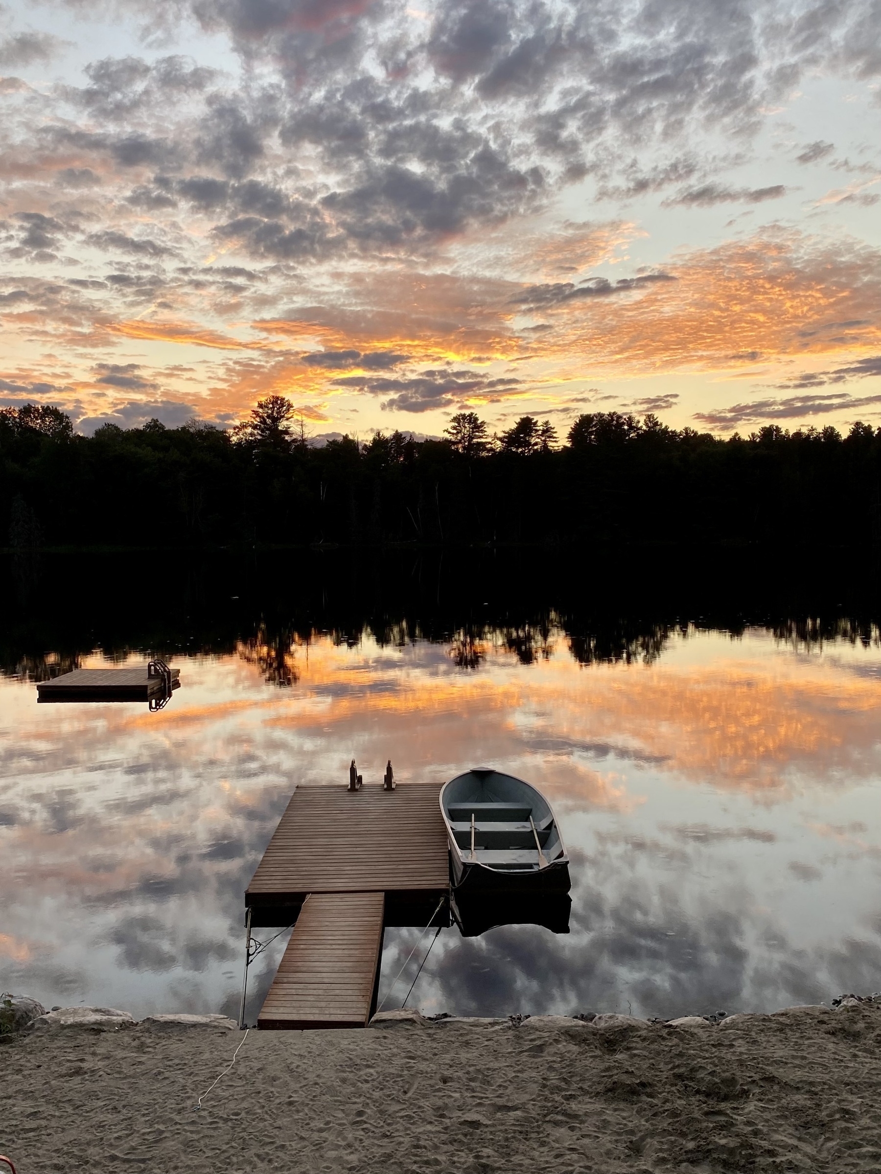 A boat docked on a serene lake reflects the cloudy sunset sky, surrounded by tranquil trees and a sandy shore.