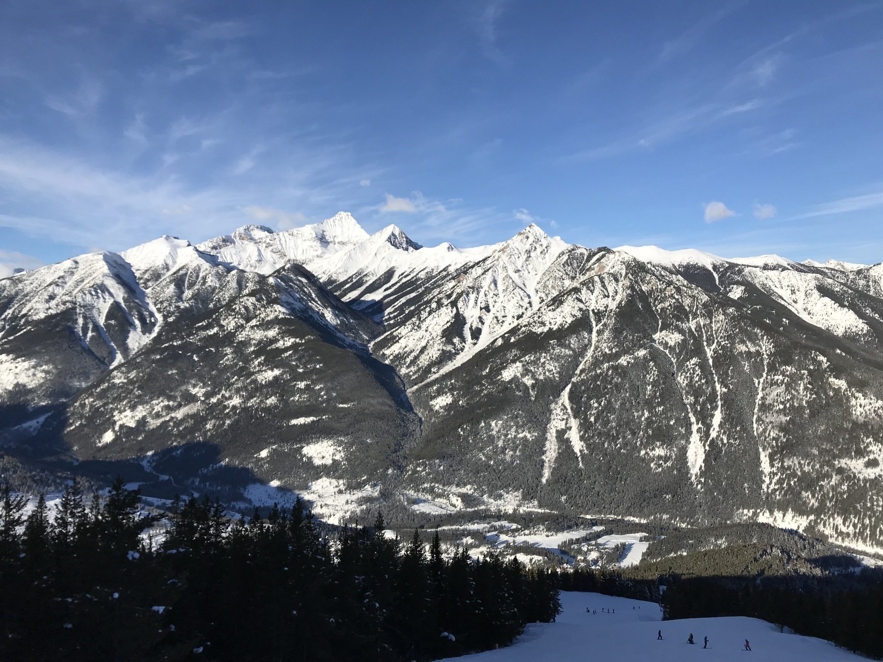 蓝天下白雪覆盖的山脉，前景是在广阔崎岖的高山景观中滑雪的小人物。