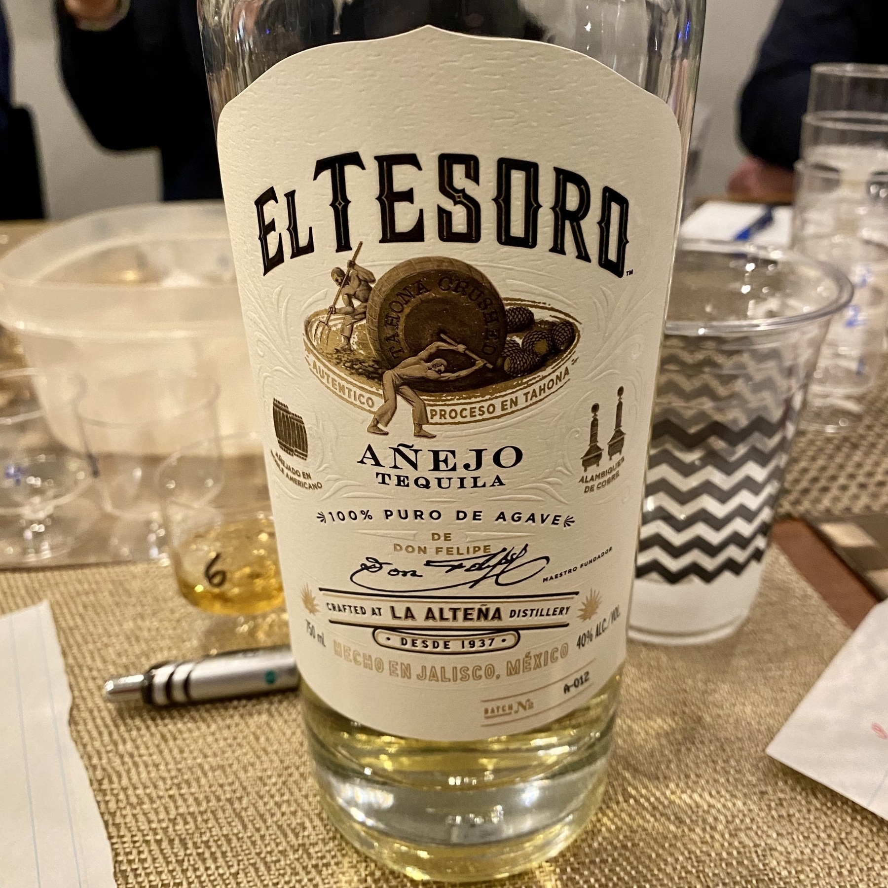 A bottle of “El Tesoro Añejo Tequila” sits on a table, featuring labels with text indicating a traditional process and a crafted distillery background.