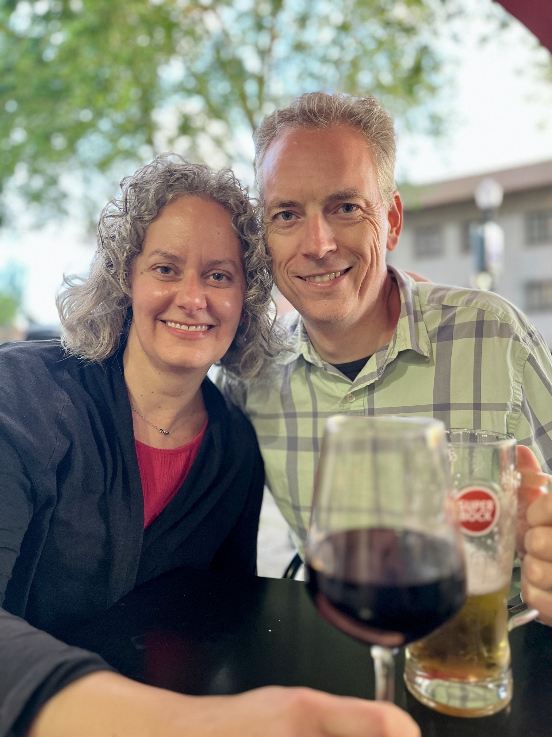 A smiling man and woman pose closely, with glasses of wine and beer on a table in front, at an outdoor seating area.