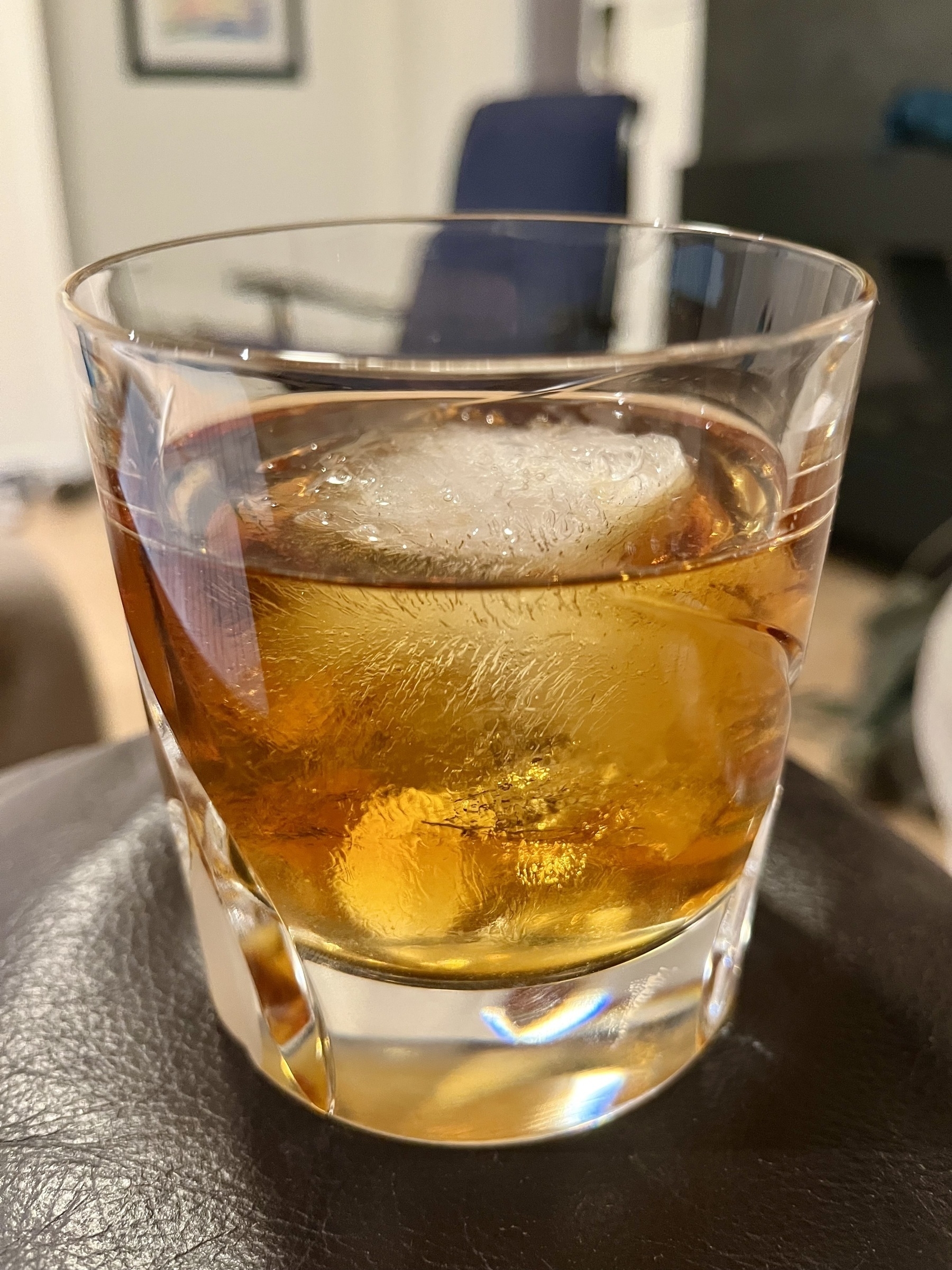 A whiskey glass holding amber liquid and a large ice cube, resting on a dark surface with a blurred indoor background.