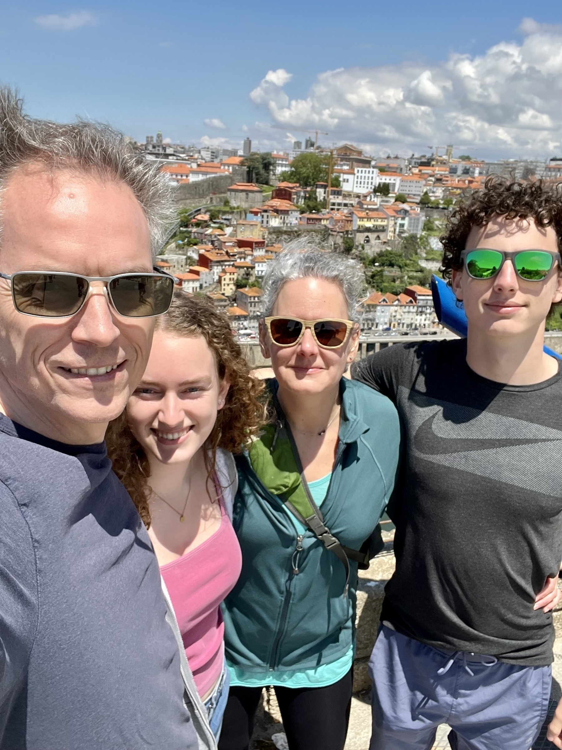 Four people are posing for a selfie with a scenic city backdrop under a cloudy sky. They are smiling and wearing casual attire with sunglasses.