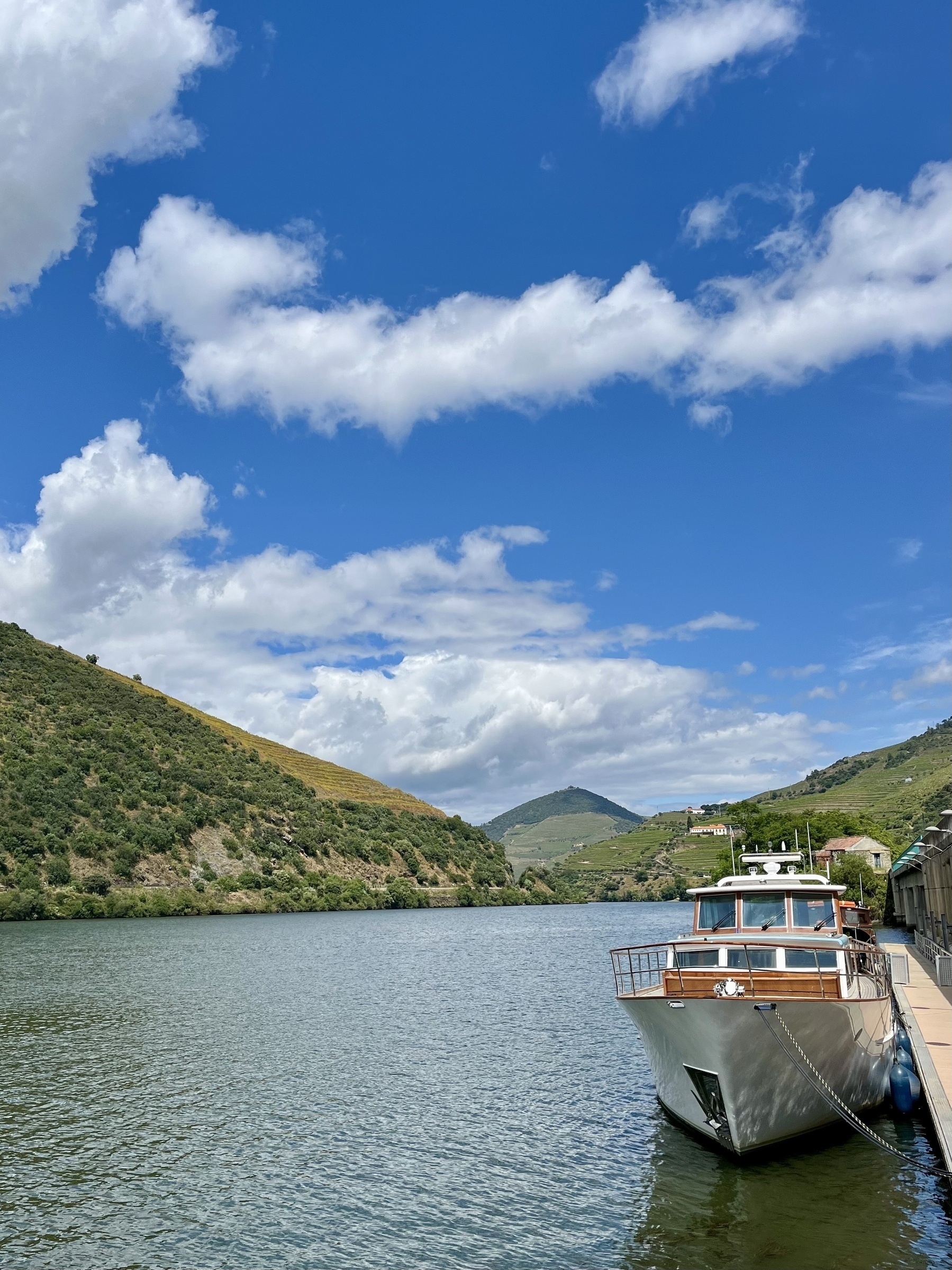 A boat moored by a river, with green hills in the background under a blue sky dotted with fluffy clouds.
