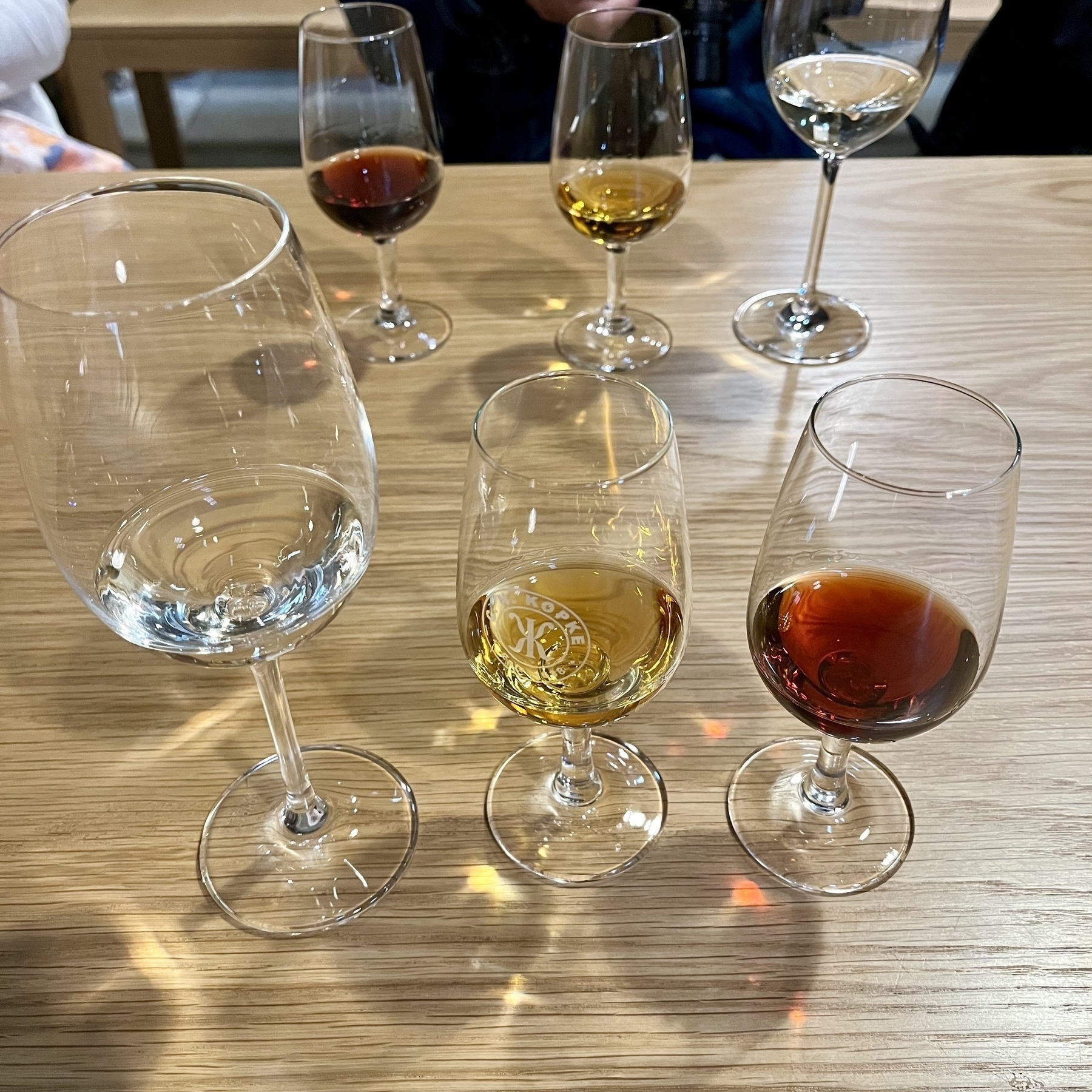 Five glasses rest on a table, containing clear, golden, and dark liquids, suggesting a tasting event. The wooden surface and glassware convey a social setting.