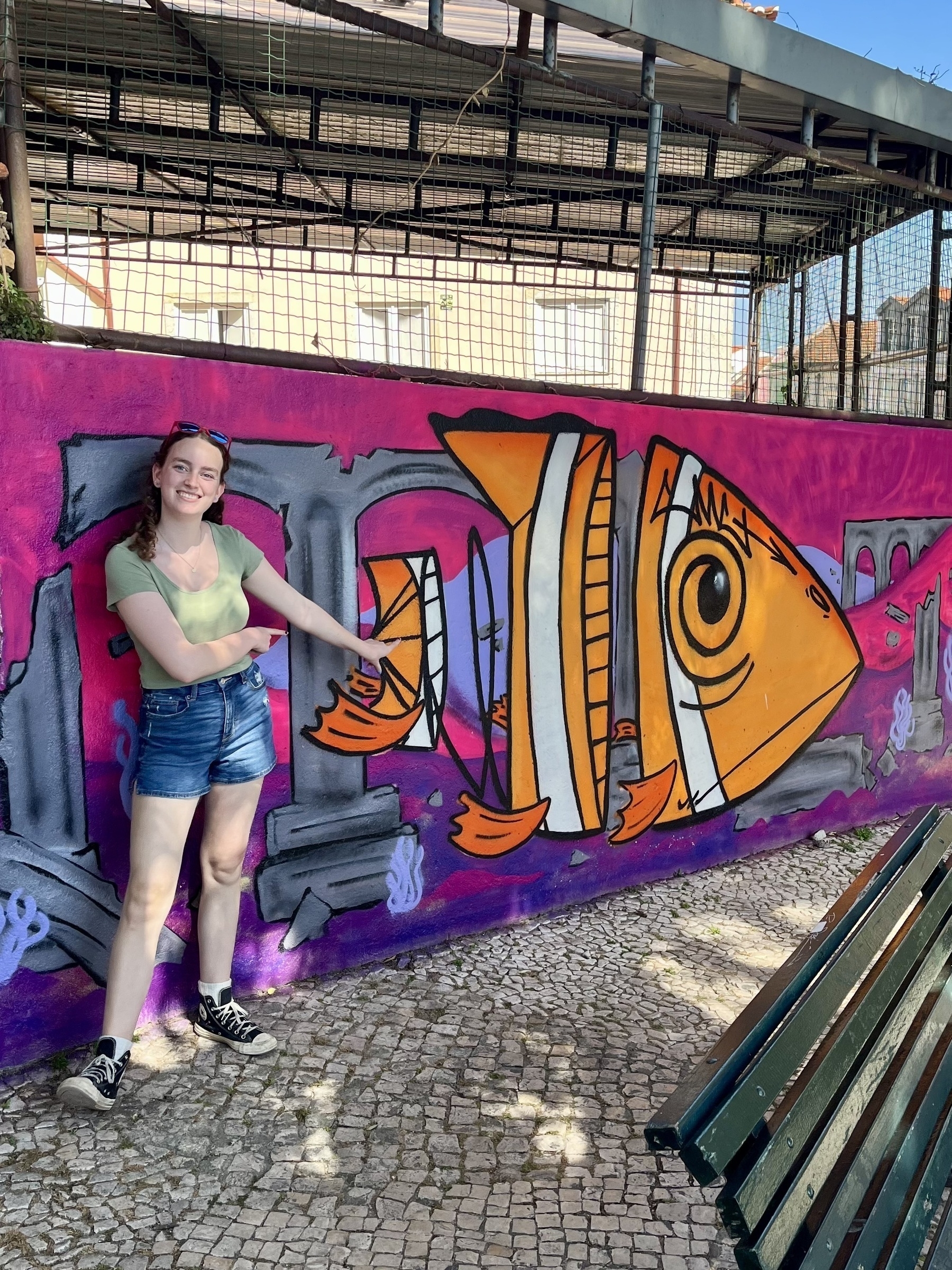 A smiling person leans against a colorful mural of a fish; urban setting with cobblestones and benches.