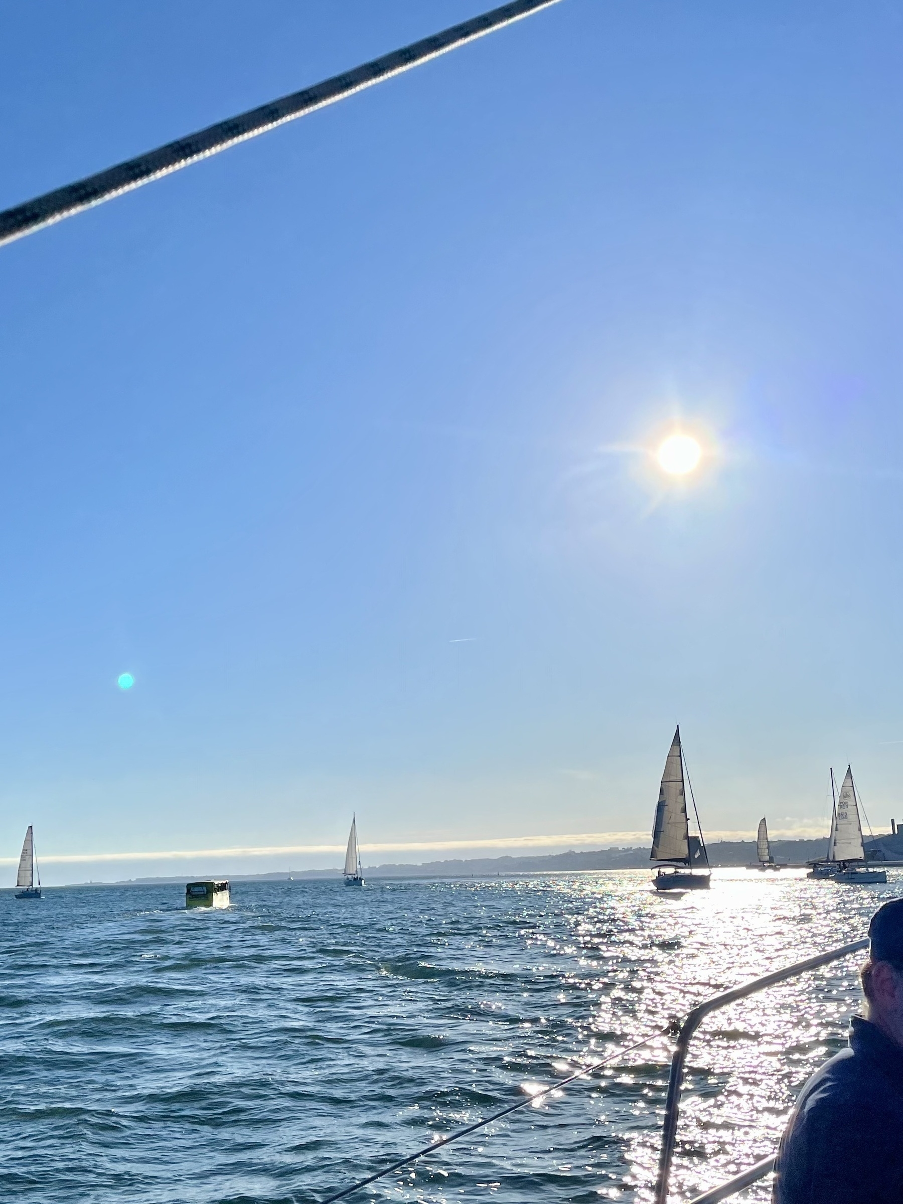 Sailboats glide across glittering water with a sun low in the blue sky, viewed from a boat with a visible cable and part of a person's head.