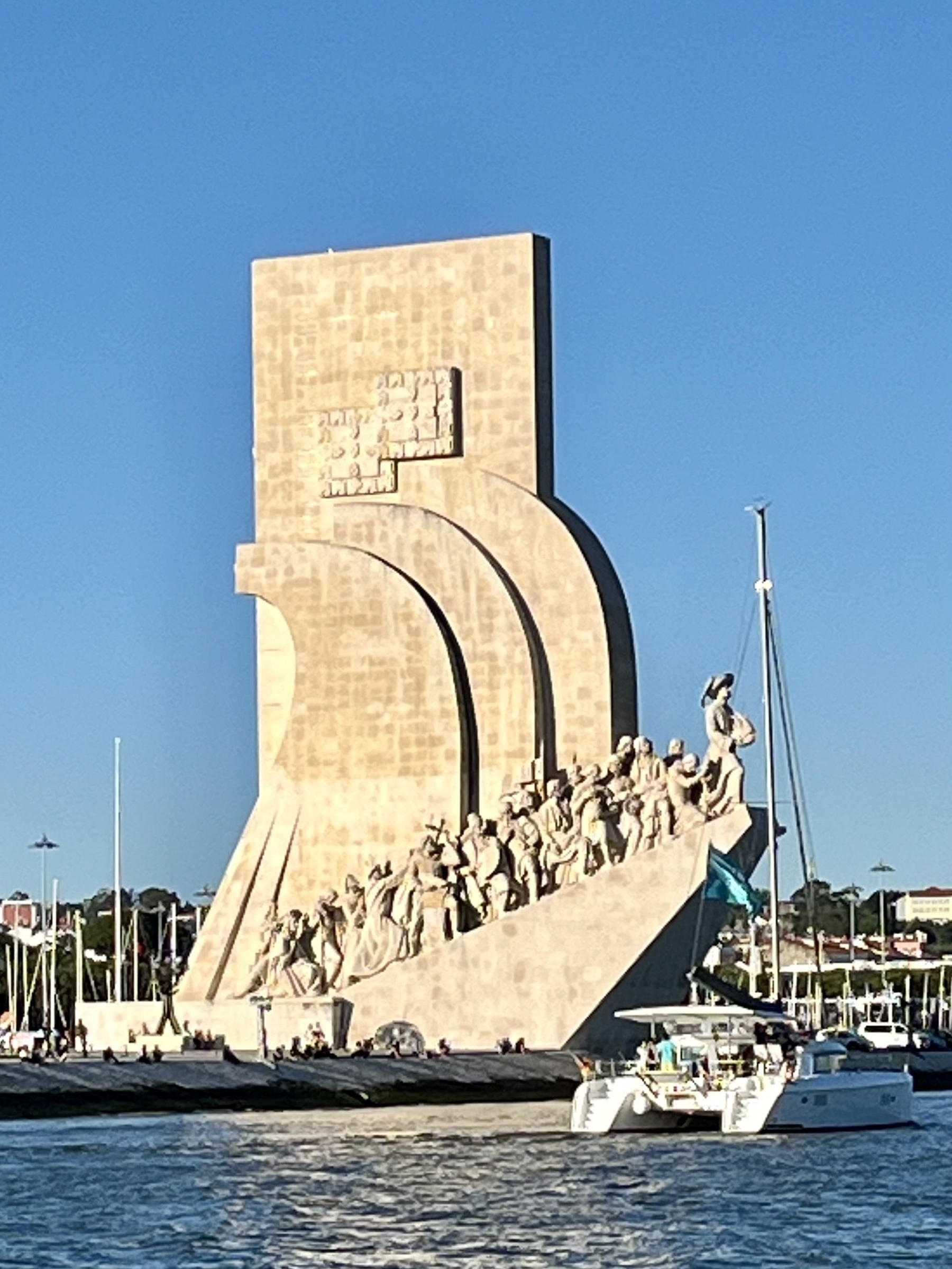 A monument featuring sculptural figures ascending along its edge, stands beside a marina with boats, under a clear blue sky.